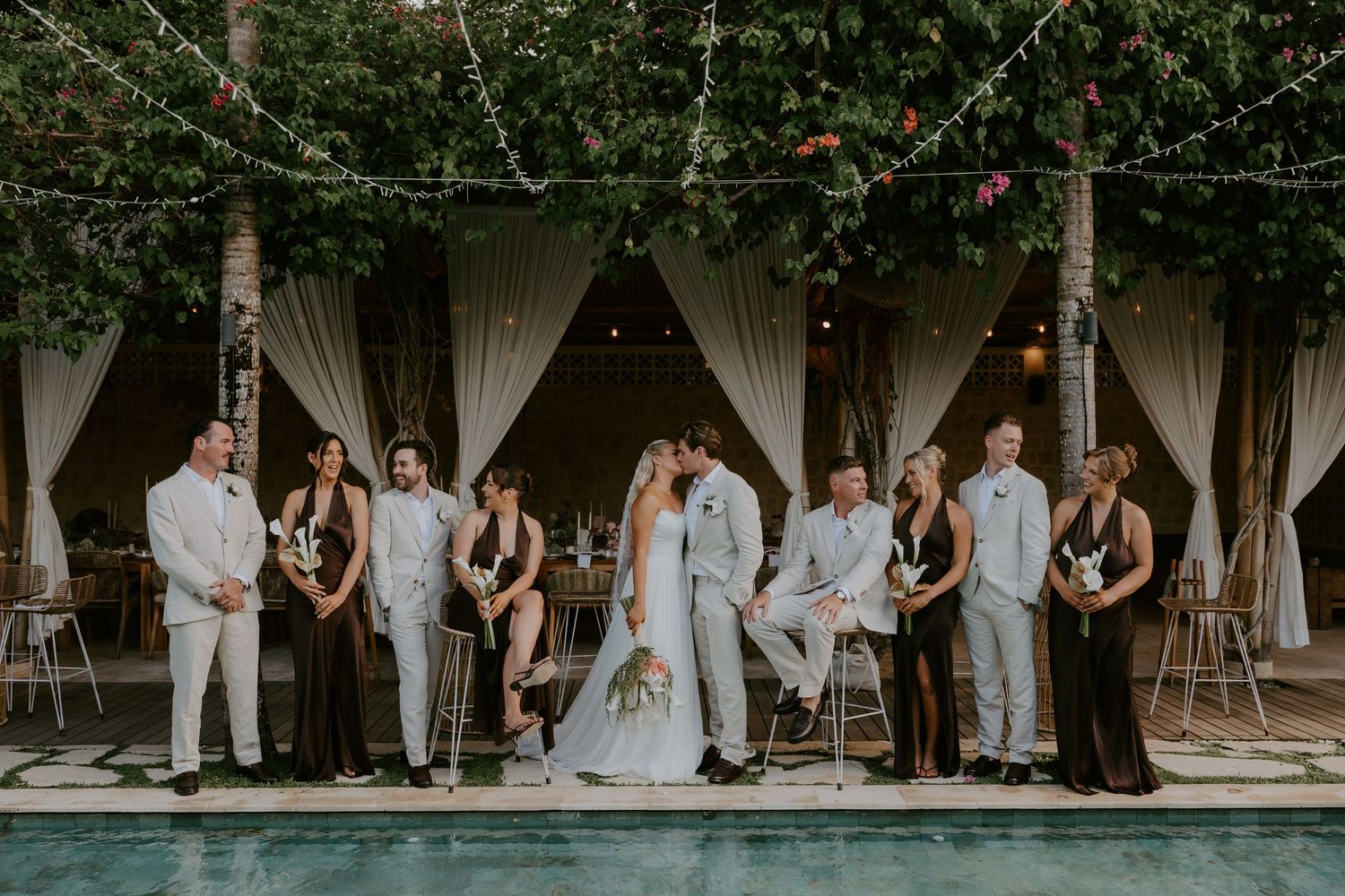 Wedding party poses poolside under string lights and lush greenery, bride and groom kissing center.