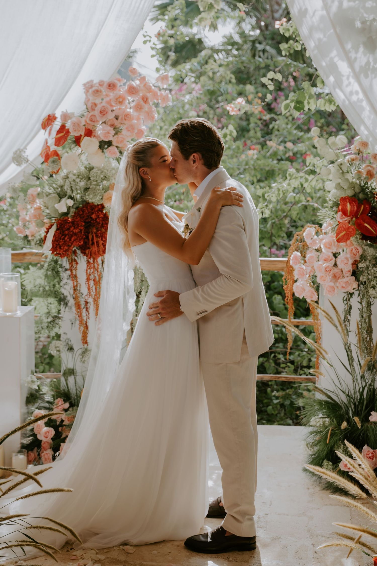 Bride and groom share first kiss under floral arch with pink and red blooms at outdoor wedding.