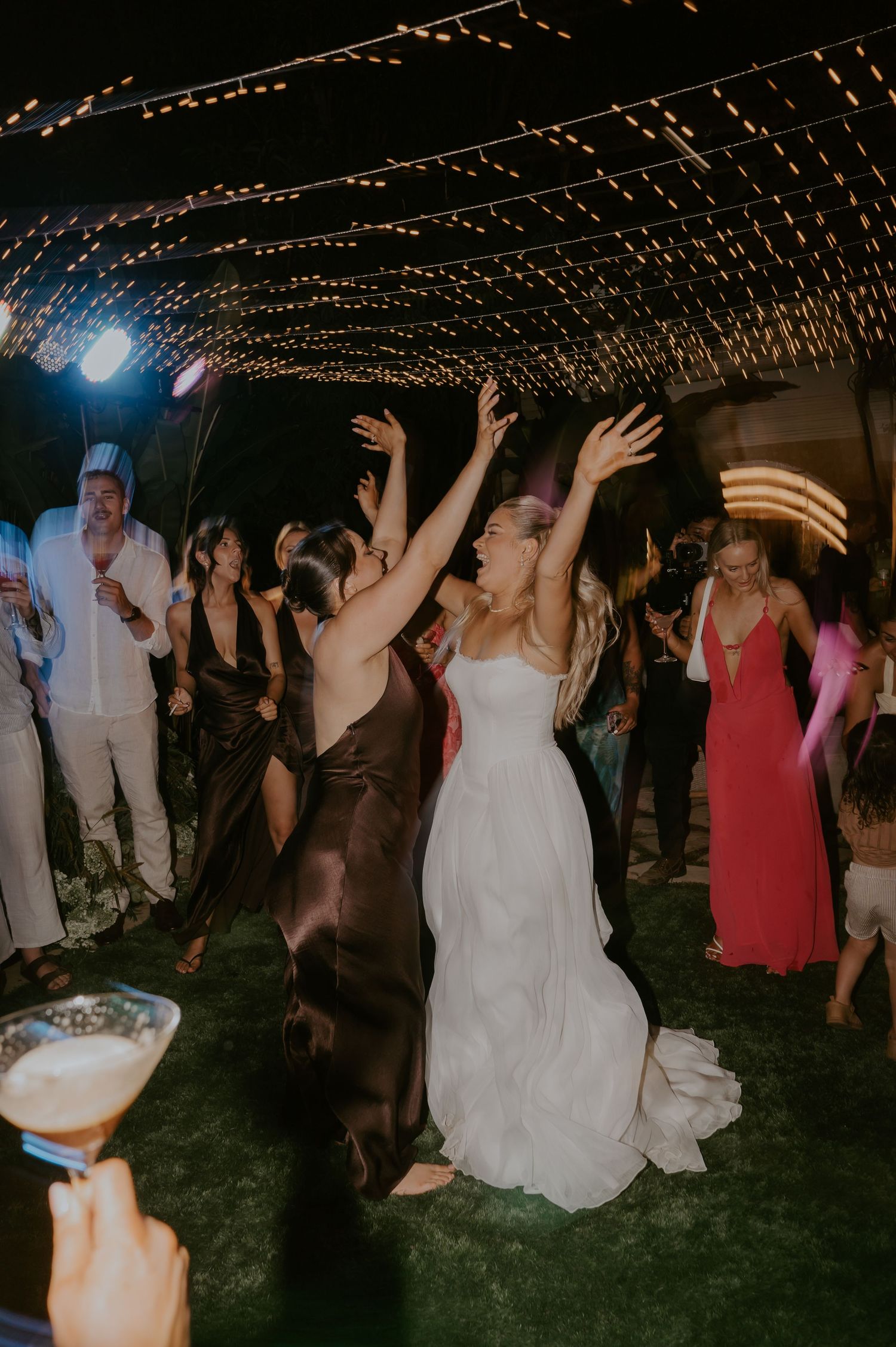 Bride and guests dance with arms raised under string lights at night reception.