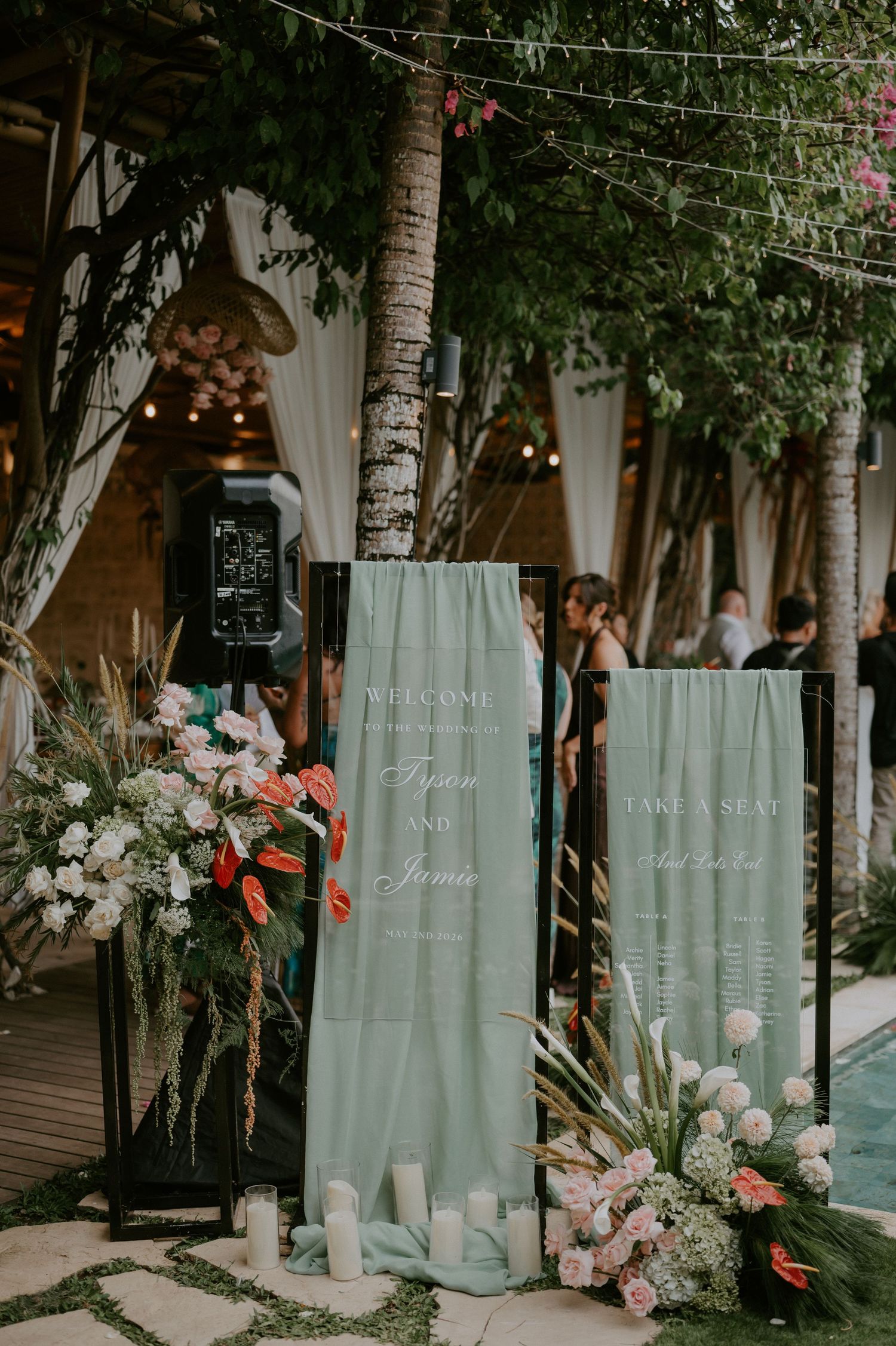 Elegant sage green wedding welcome signage with tropical floral arrangements and candles at outdoor venue.