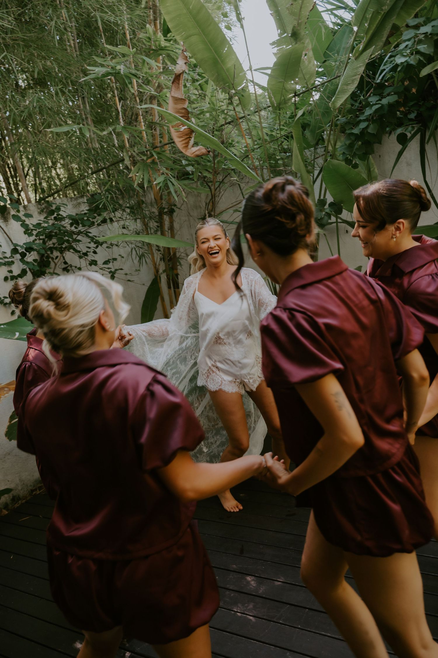 Bride laughing with bridesmaids in burgundy robes on a tropical wooden deck surrounded by lush greenery.
