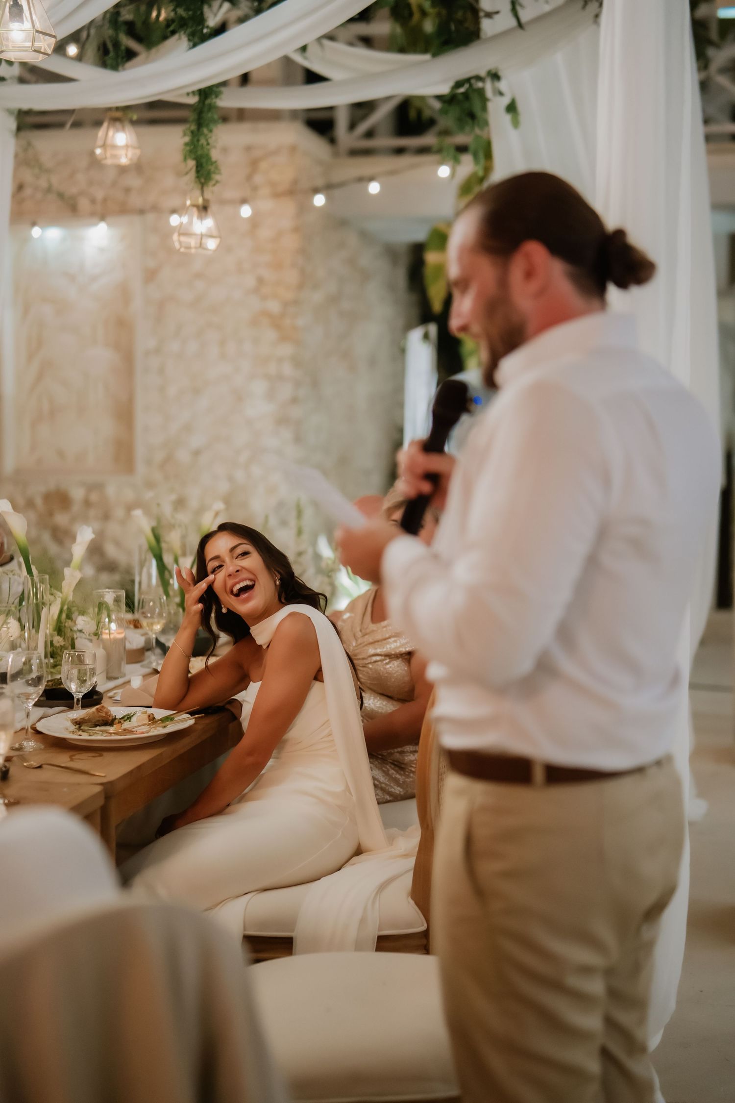 Bride laughs joyfully as groom delivers a speech at a candlelit wedding reception with draped decor.