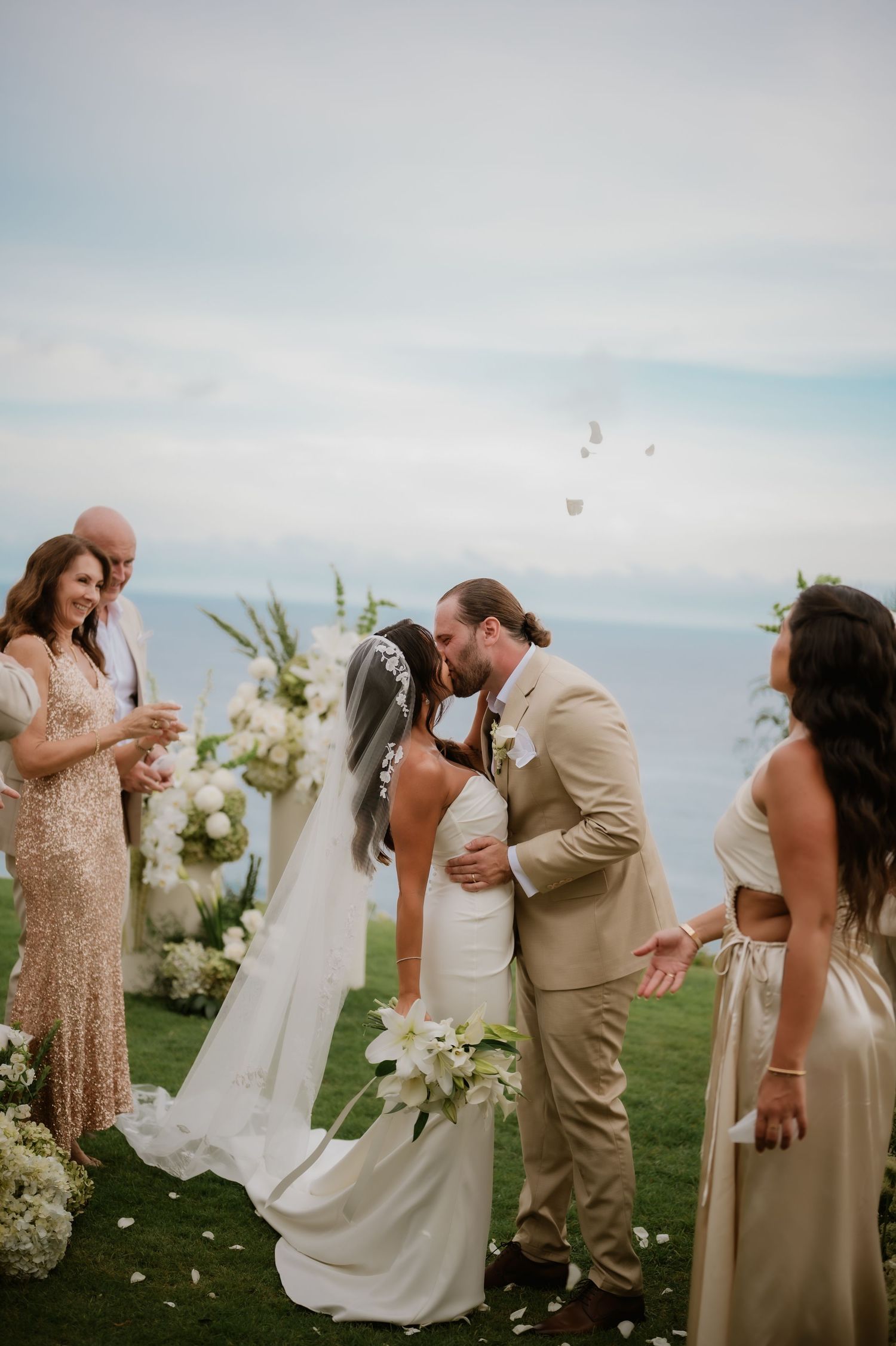 Bride and groom share first kiss at outdoor wedding ceremony while guests celebrate with petals.