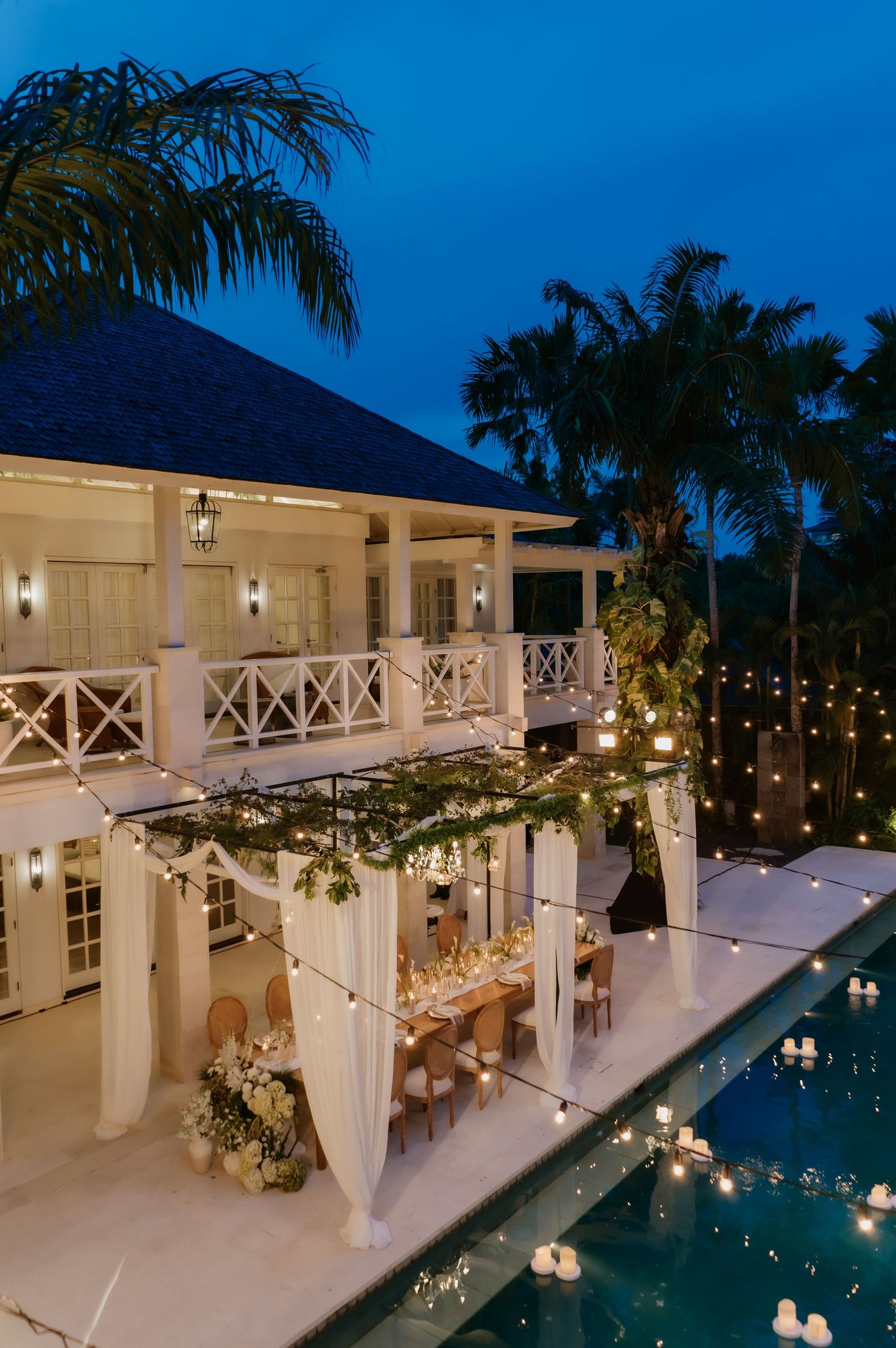 Elegant outdoor wedding reception beside pool with string lights, draped canopy, and tropical villa at dusk.