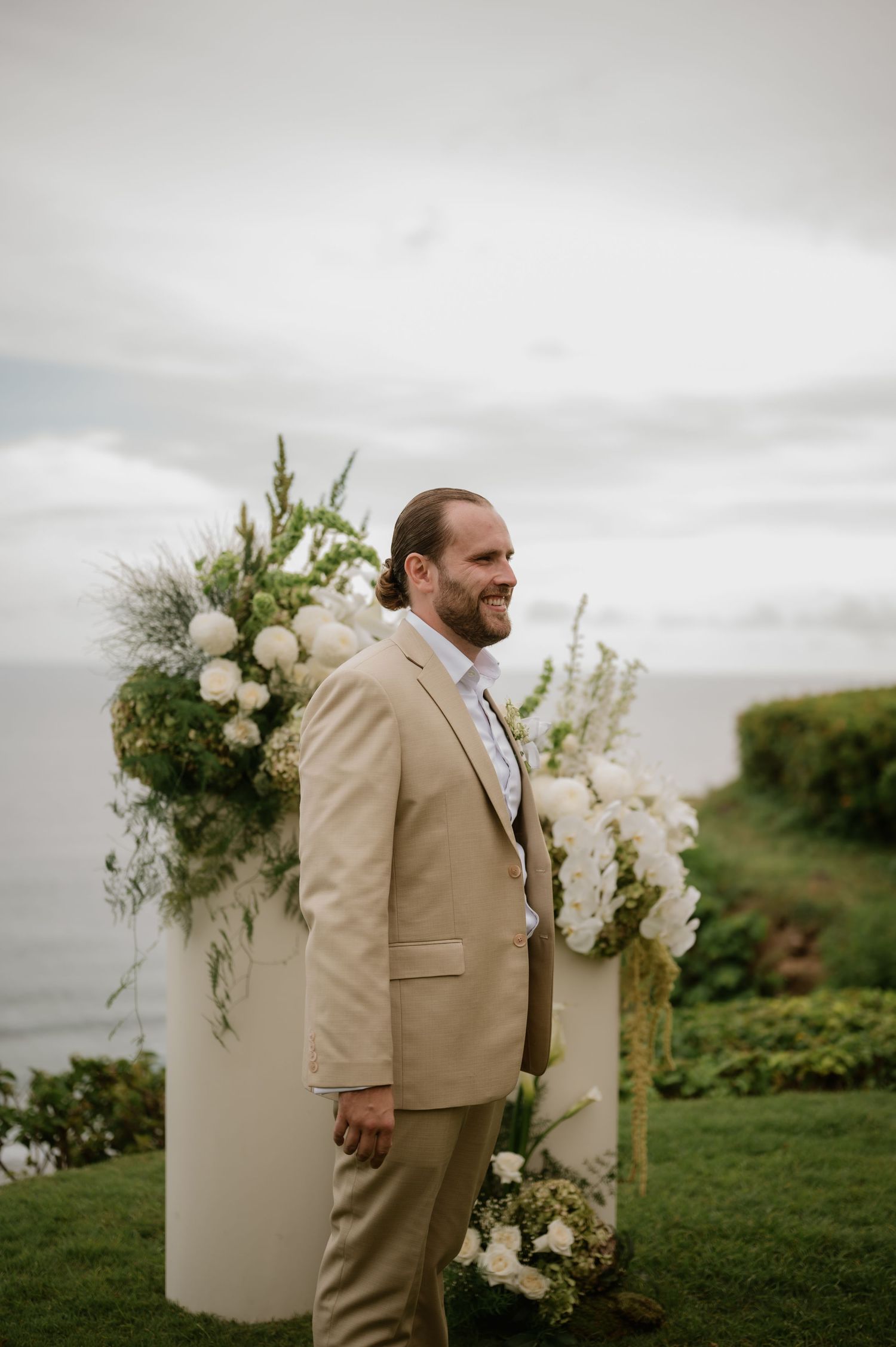 Groom in tan suit smiling at outdoor wedding ceremony with white floral arch and ocean view.