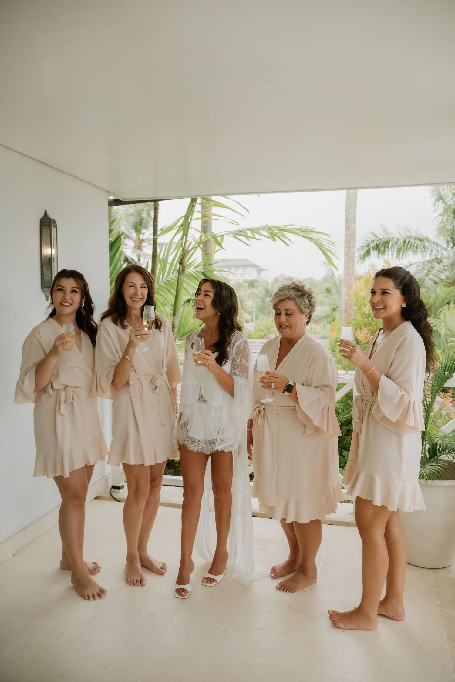 Bride and bridesmaids laughing and toasting with champagne glasses in matching cream robes at a tropical venue.