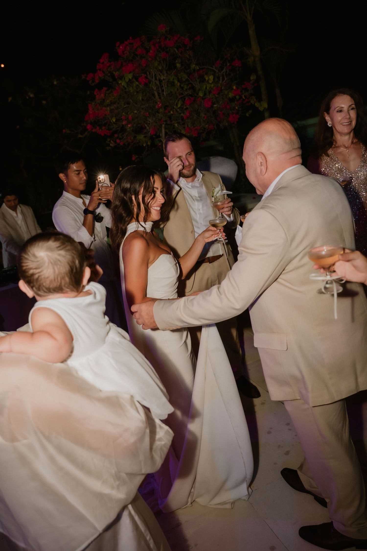 Bride and groom dancing joyfully with guests at lively outdoor tropical wedding reception at night.