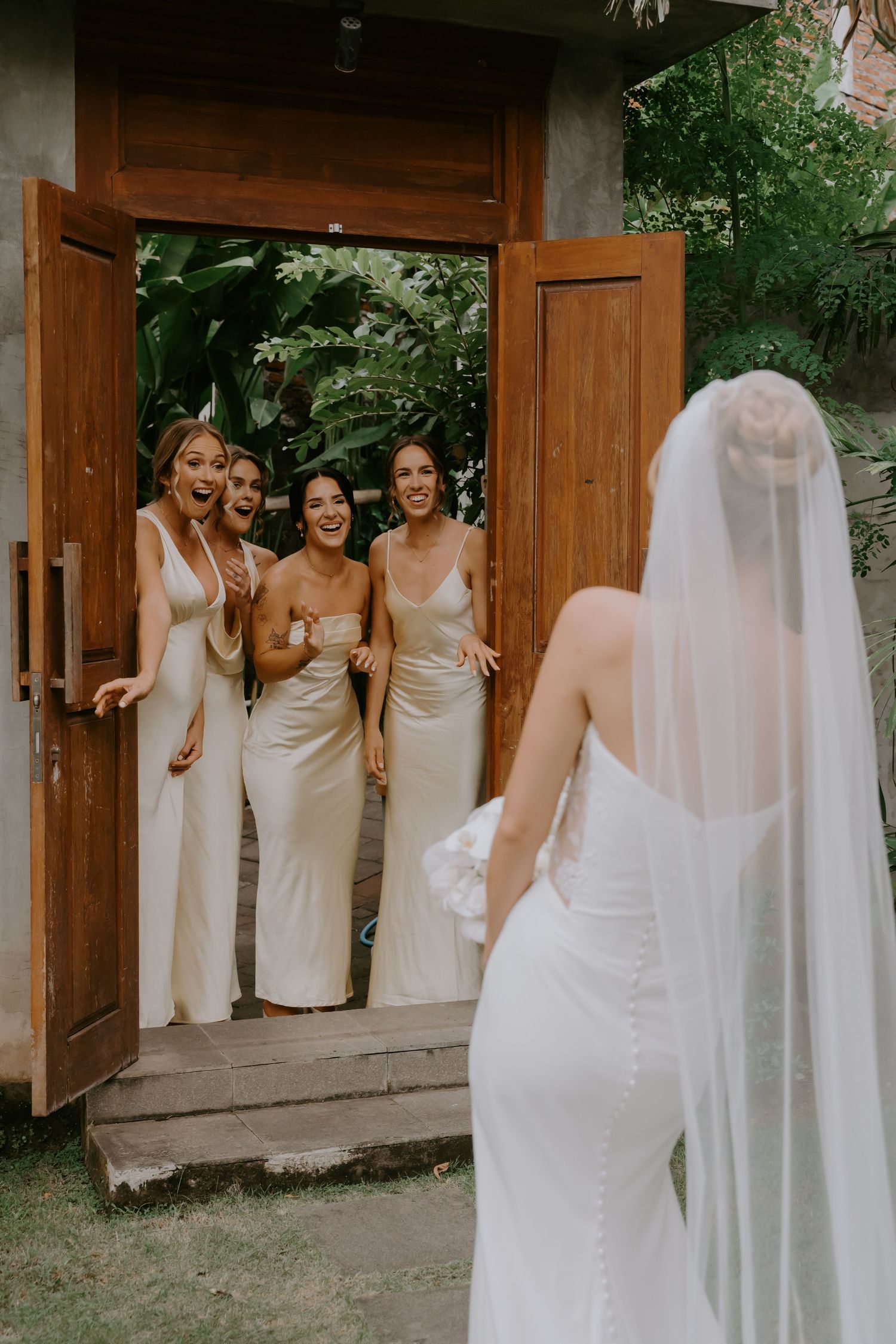Bridesmaids in champagne satin gowns react with joy seeing bride in white wedding dress.