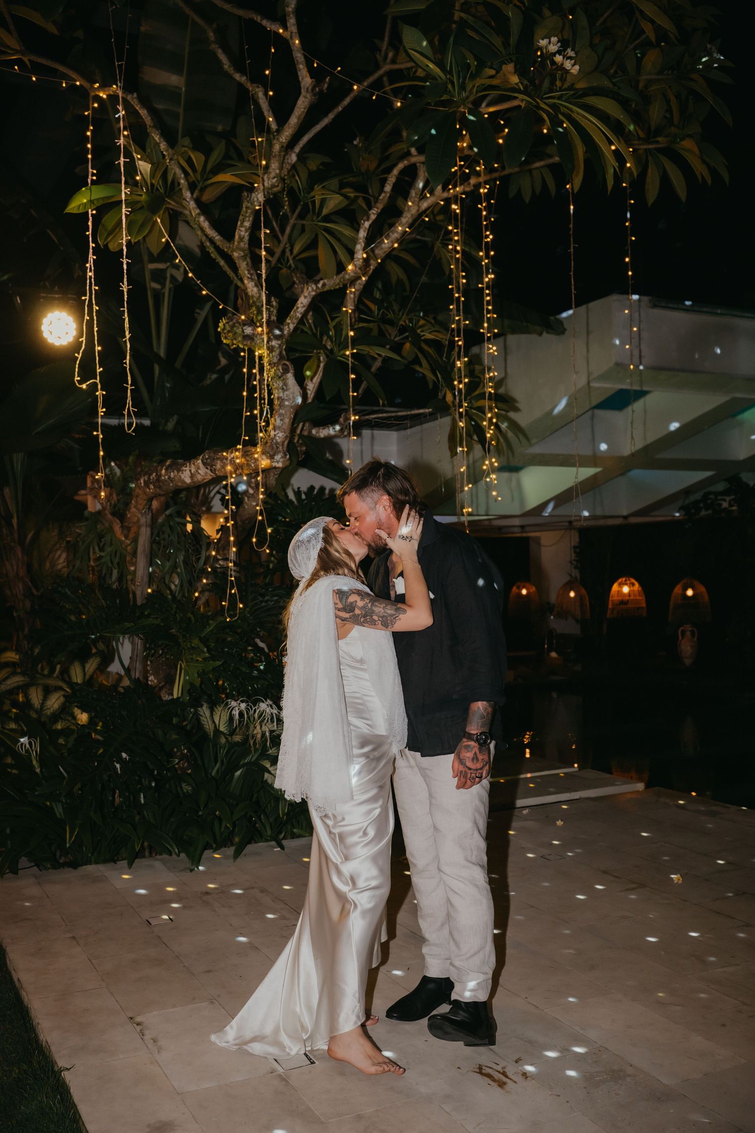 Newlyweds share a romantic kiss under fairy light-draped trees during outdoor evening reception.