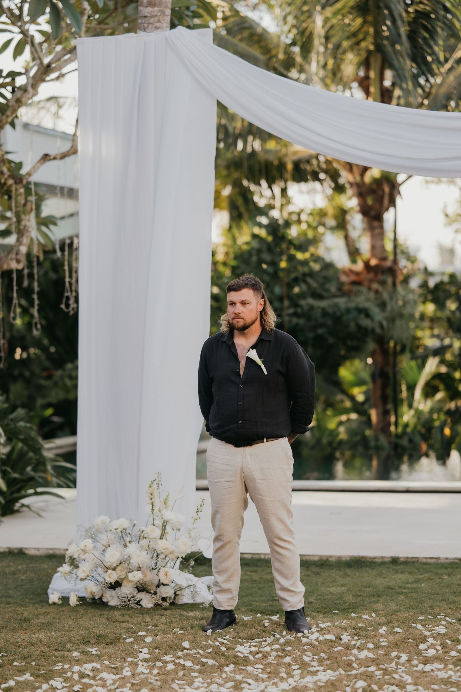Groom in black shirt and beige pants stands at outdoor wedding altar with white draping and floral arrangements.