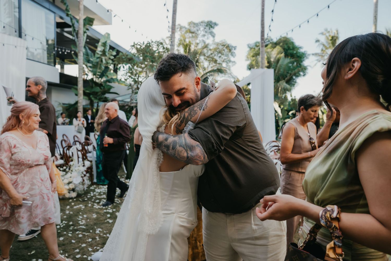 Bride in white gown and veil sharing warm embrace with tattooed guest at wedding reception.