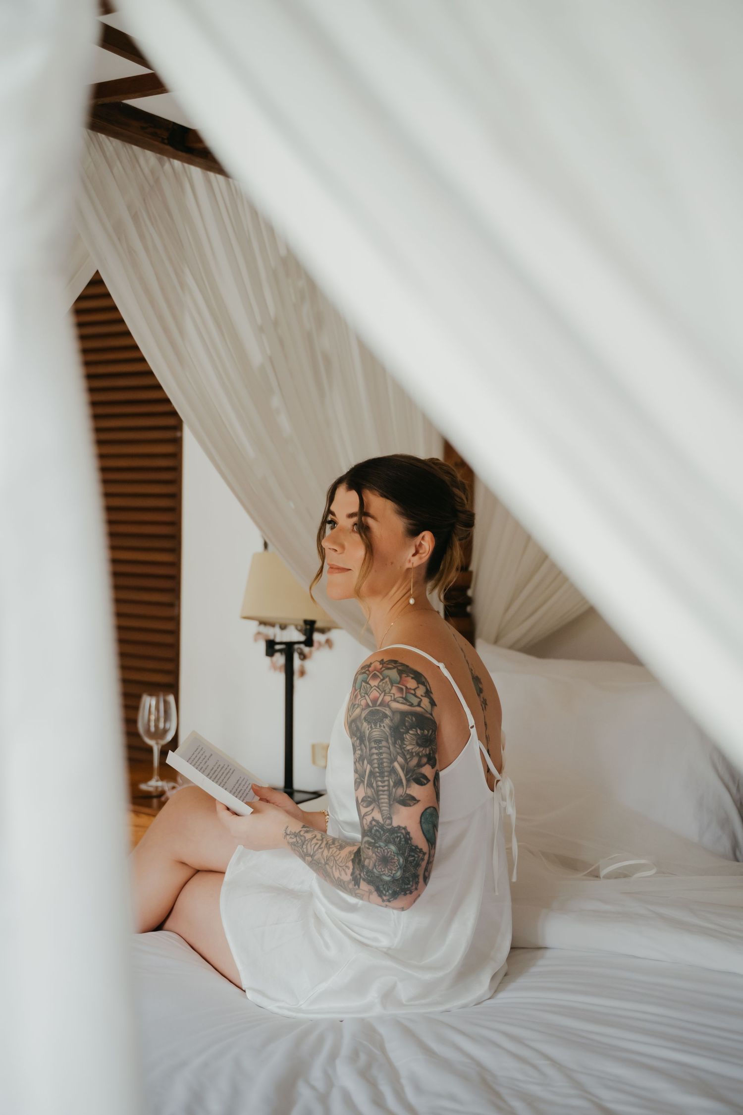 Tattooed bride in white slip dress reads on canopy bed in elegant tropical wedding villa.