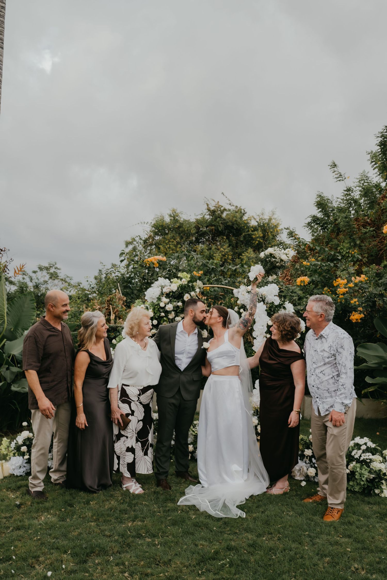 Newlyweds share a kiss with family gathered around a white floral arch in an outdoor garden ceremony.