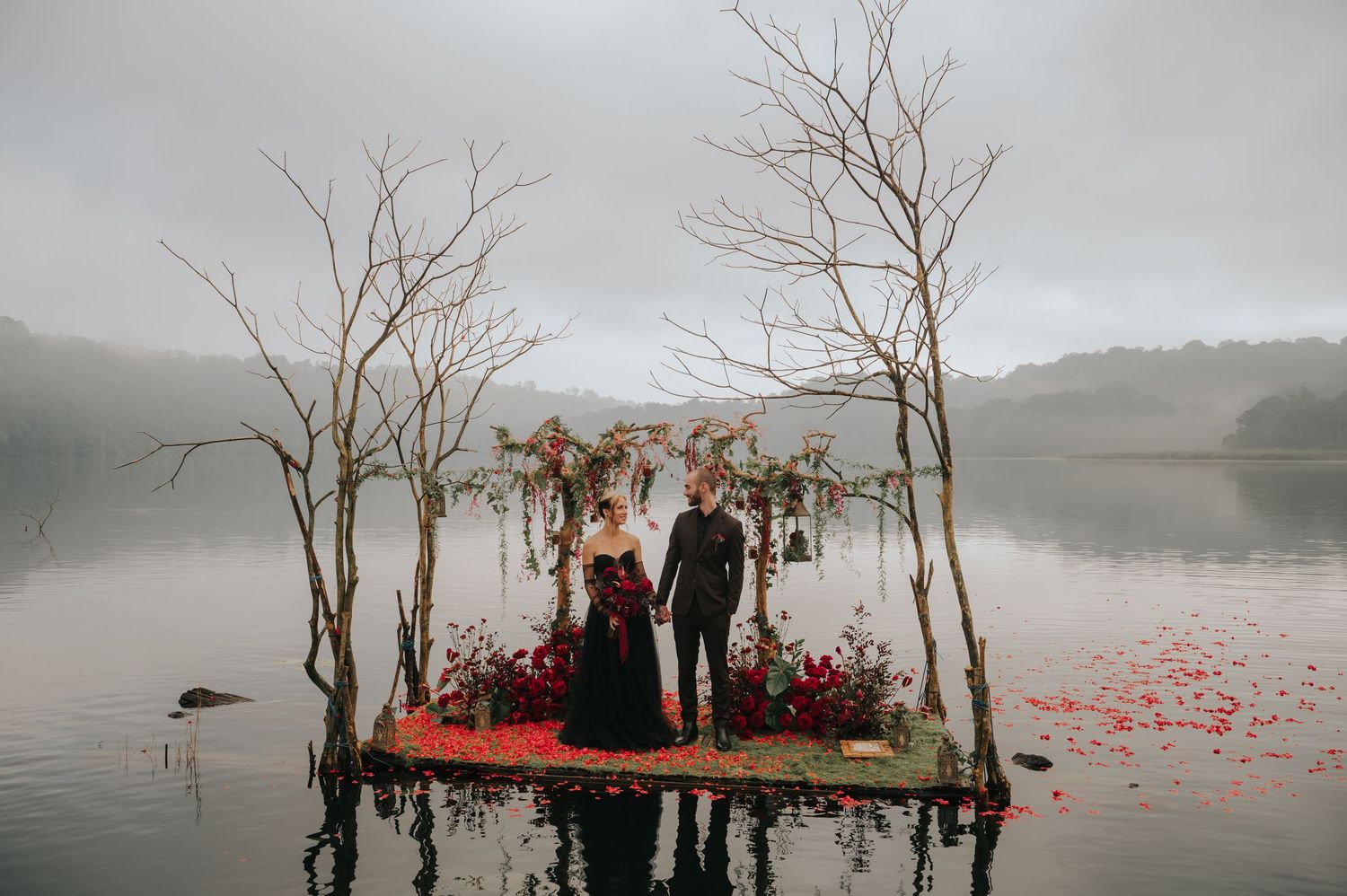 Misty lakeside wedding ceremony site with floral arches and red petals on platform.