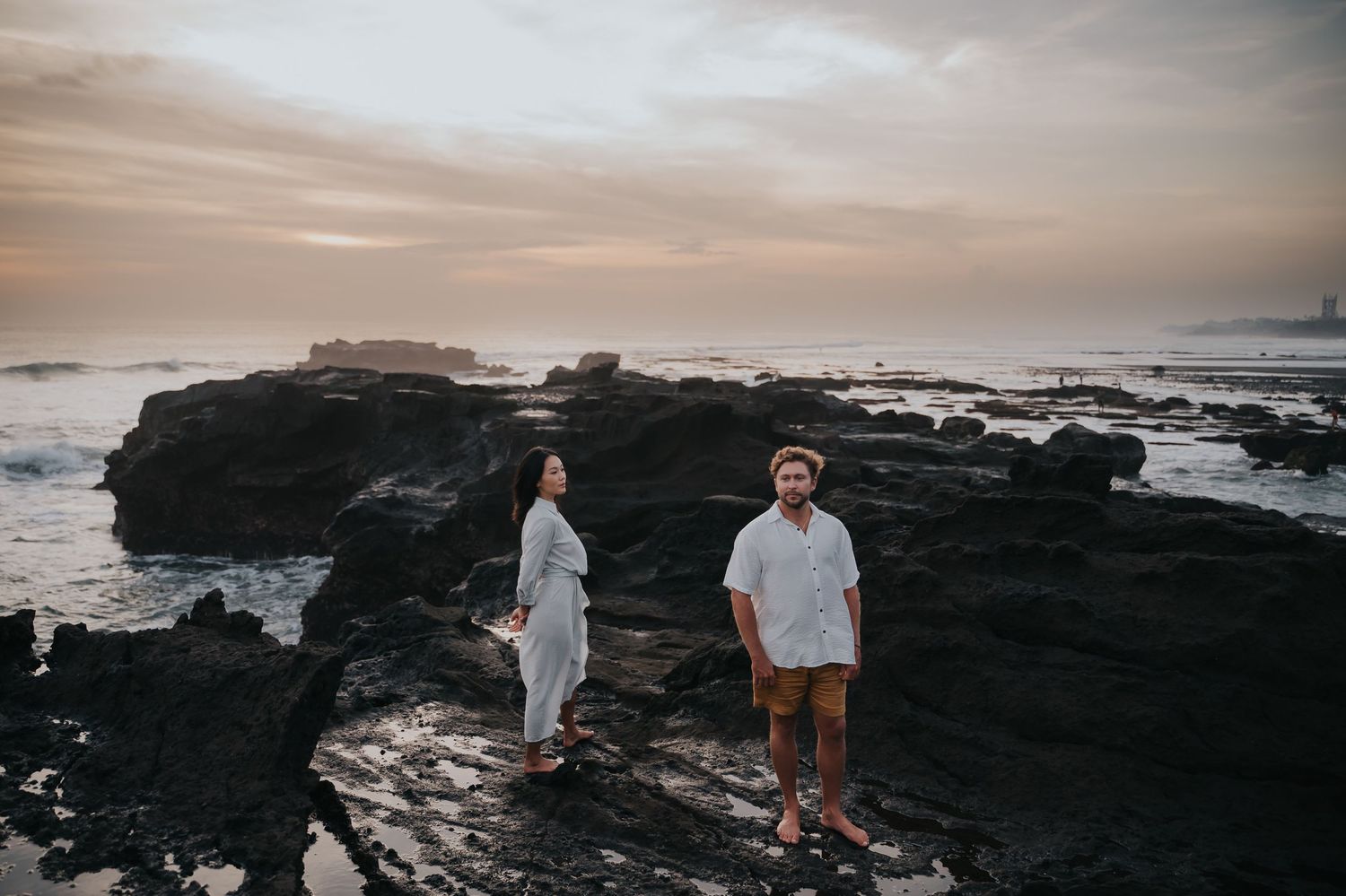 Couple walking along dramatic black volcanic rocks at sunset on a Hawaiian beach.
