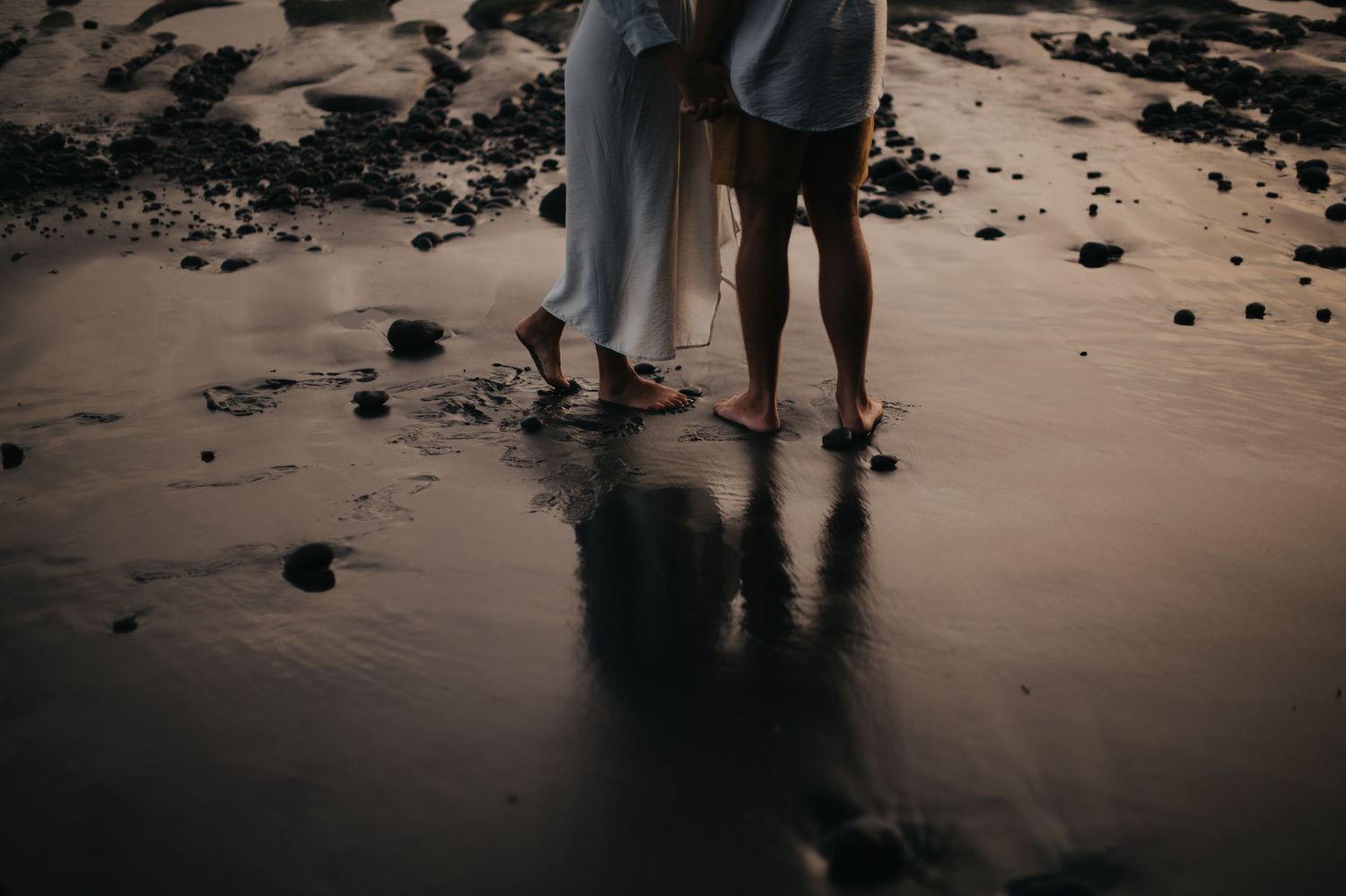 Two people standing barefoot on wet beach sand at sunset with dark shadows reflecting on the smooth surface.