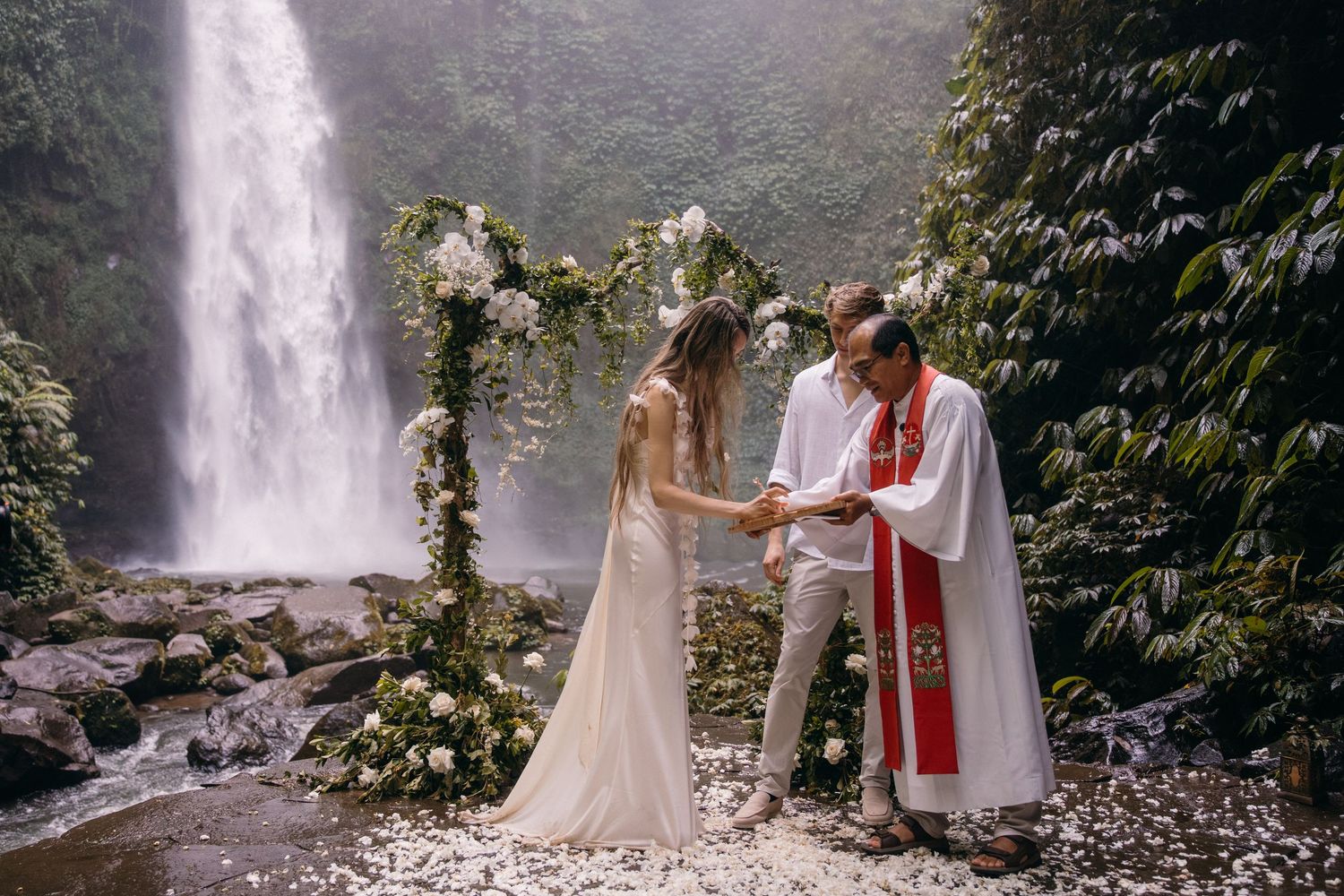 A wedding ceremony takes place in front of a majestic waterfall surrounded by lush greenery.