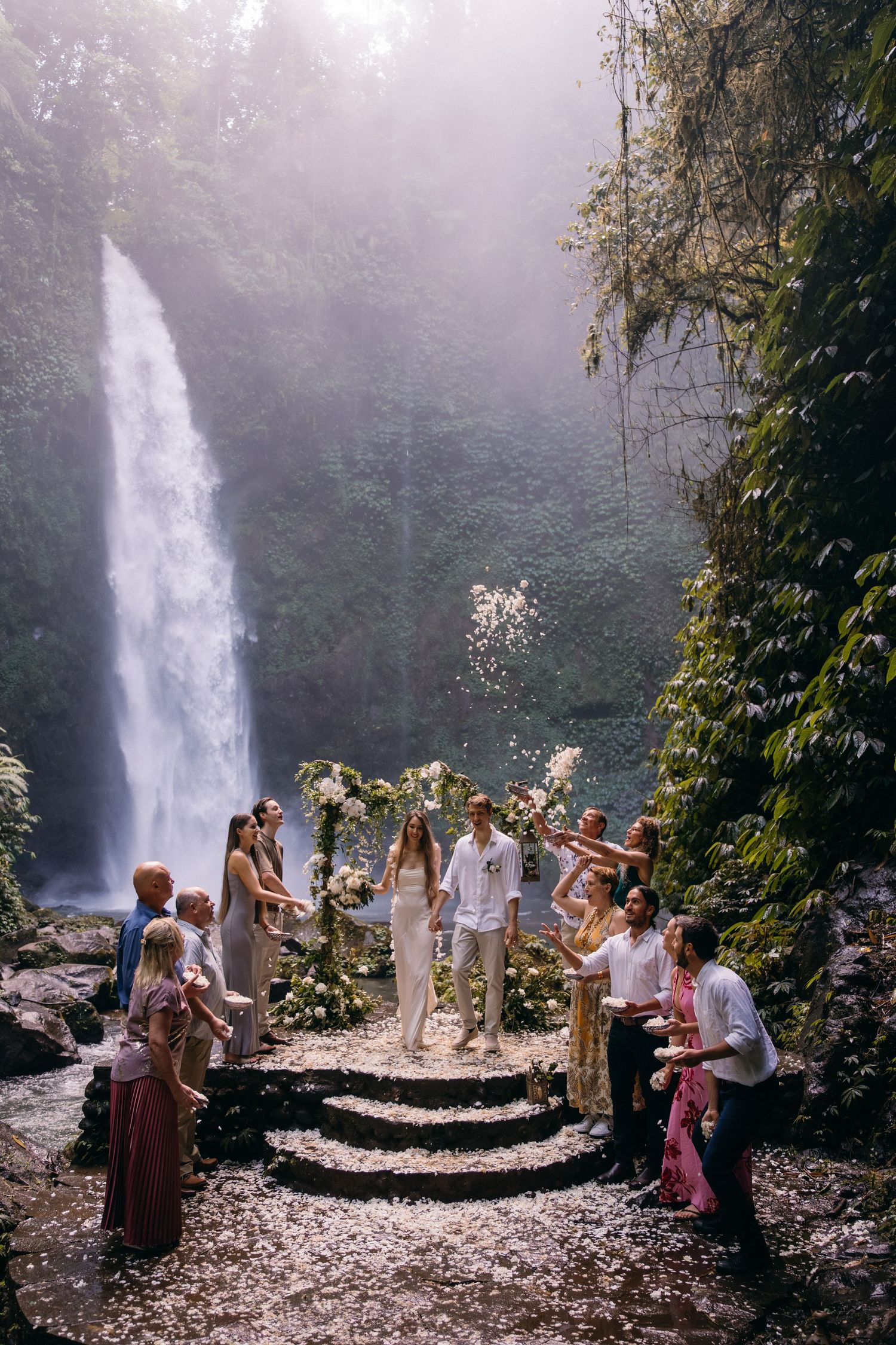 A group gathers near a majestic waterfall in a misty tropical forest setting for what appears to be a ceremony.