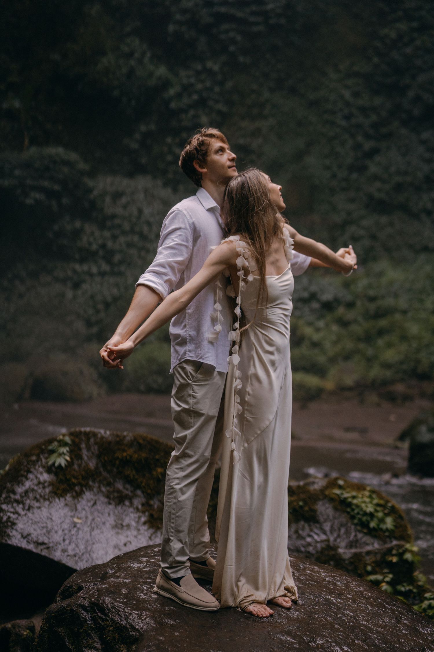 A couple in light colored outfits shares a romantic moment on a rocky cliff overlooking a scenic natural landscape.
