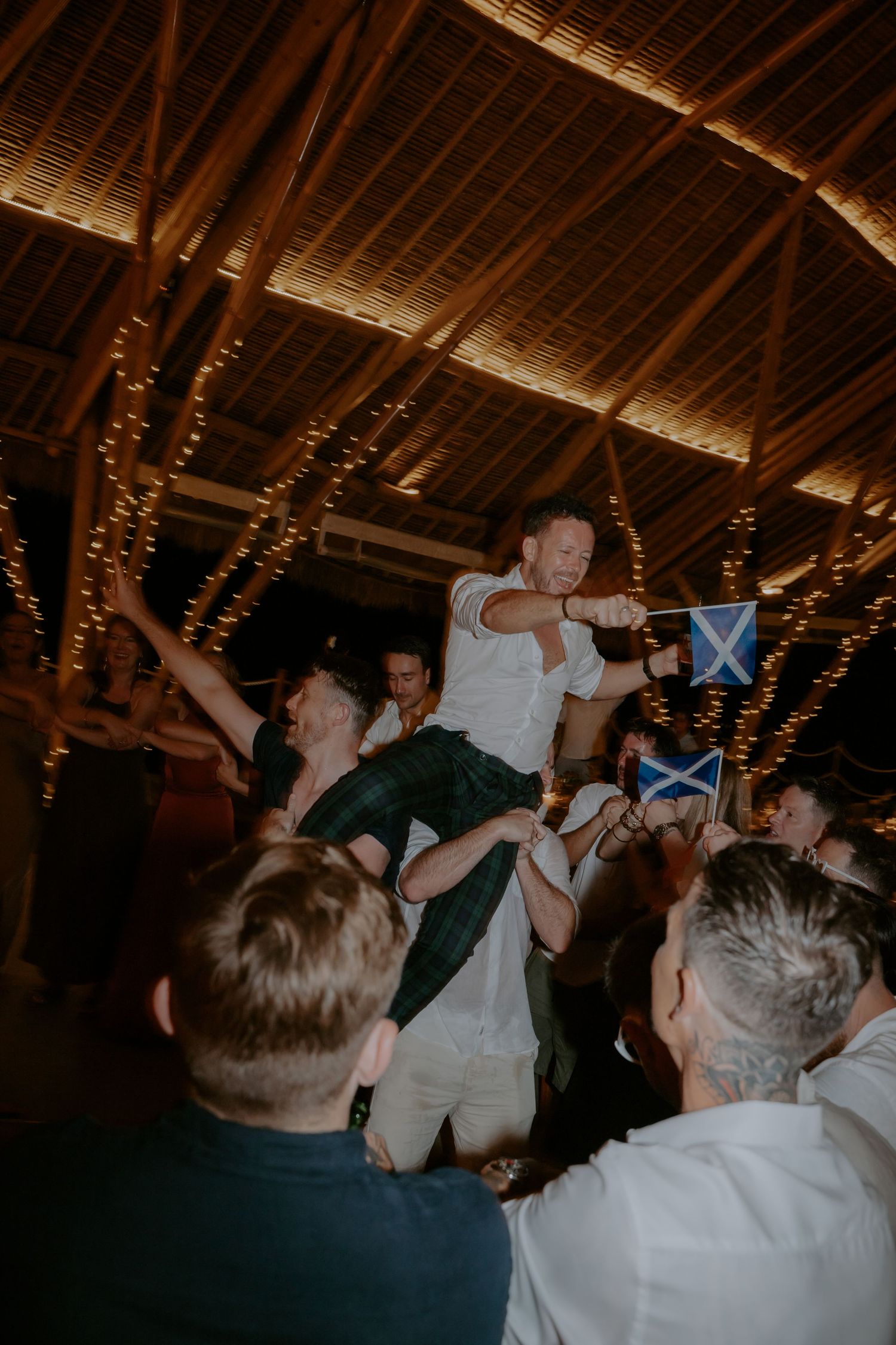 Wedding guests dance and celebrate together under rustic wooden beams decorated with string lights at a reception.