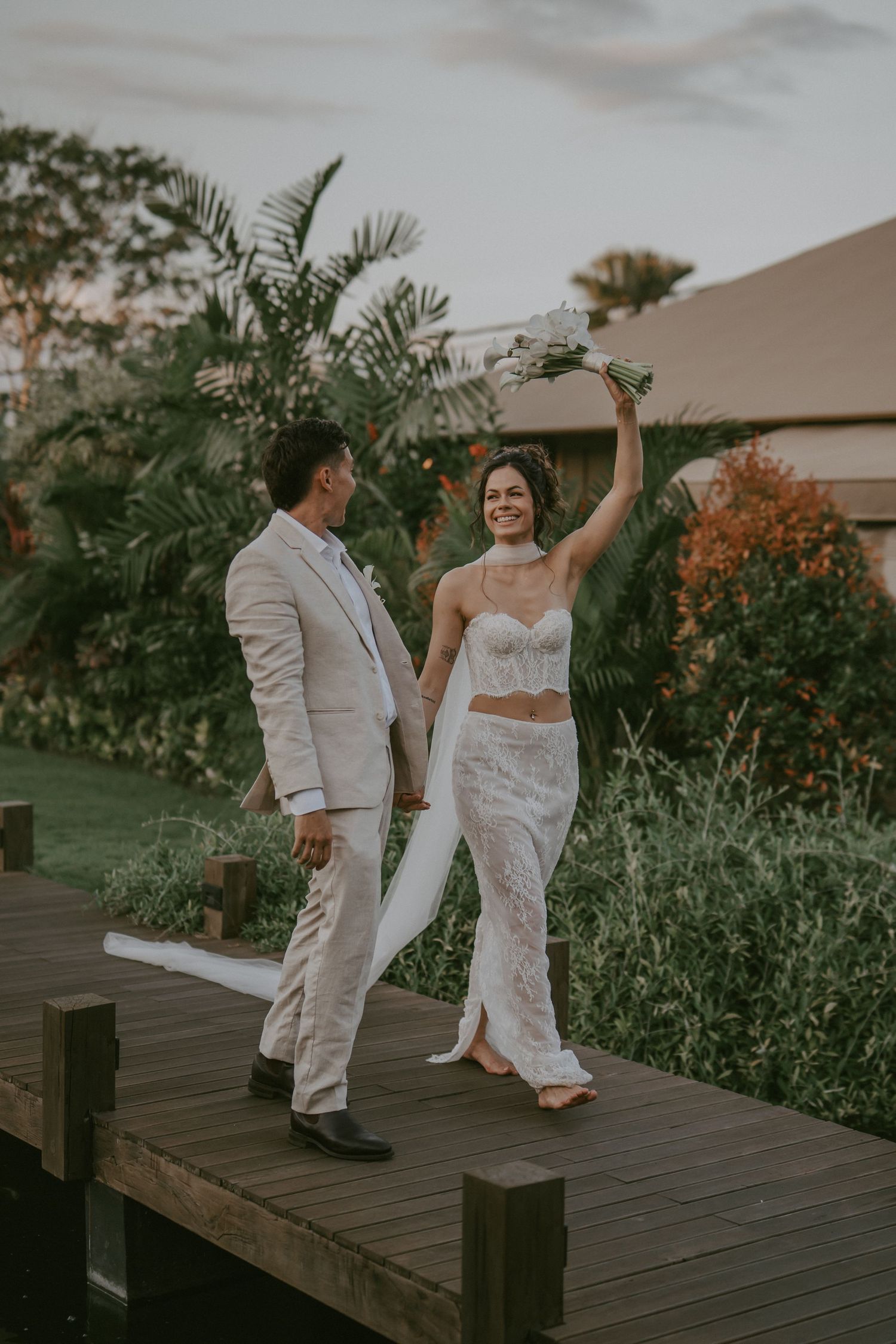 A couple in white attire dances together on a wooden deck surrounded by lush tropical gardens at sunset.