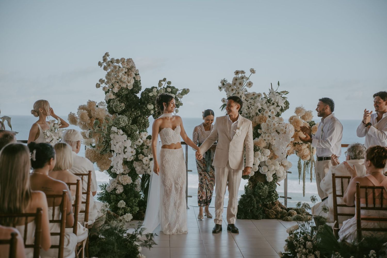 A romantic outdoor beach wedding ceremony with guests seated in white chairs and tropical floral arrangements.