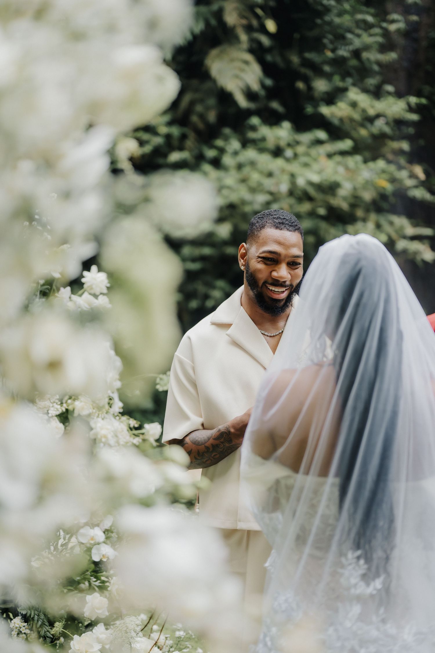 Smiling groom in cream outfit looks lovingly at veiled bride amid white wedding flowers.