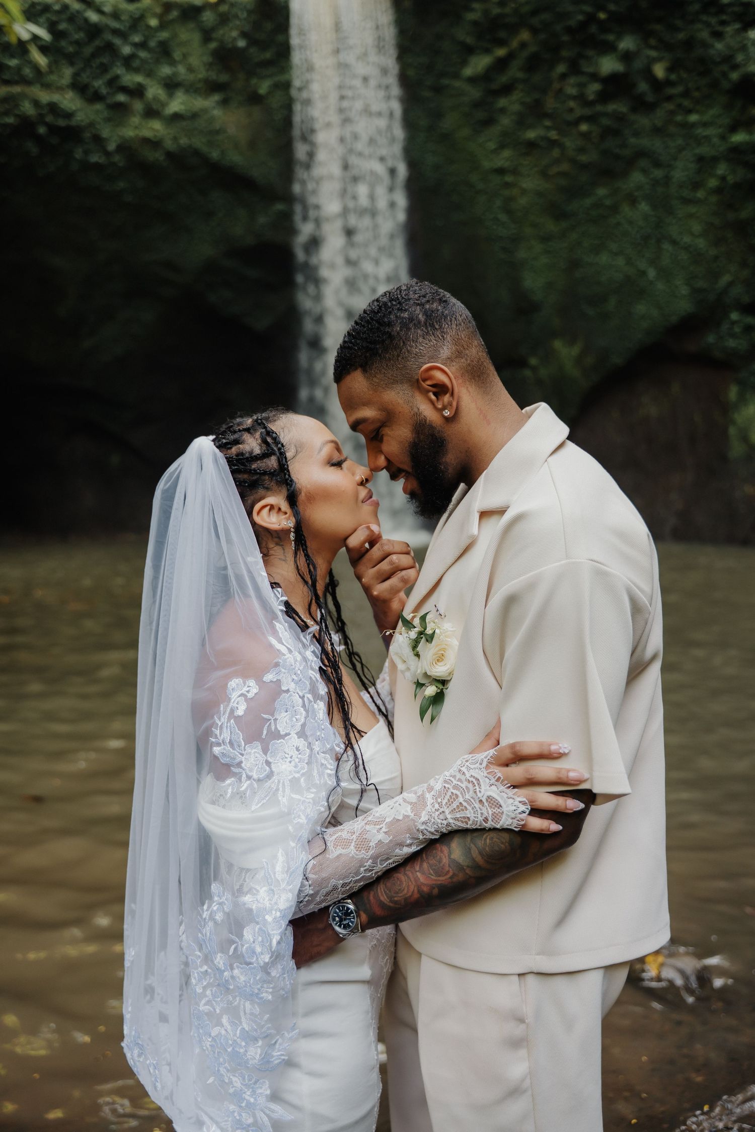 Interracial couple shares a romantic kiss near a stunning waterfall during their tropical wedding ceremony.