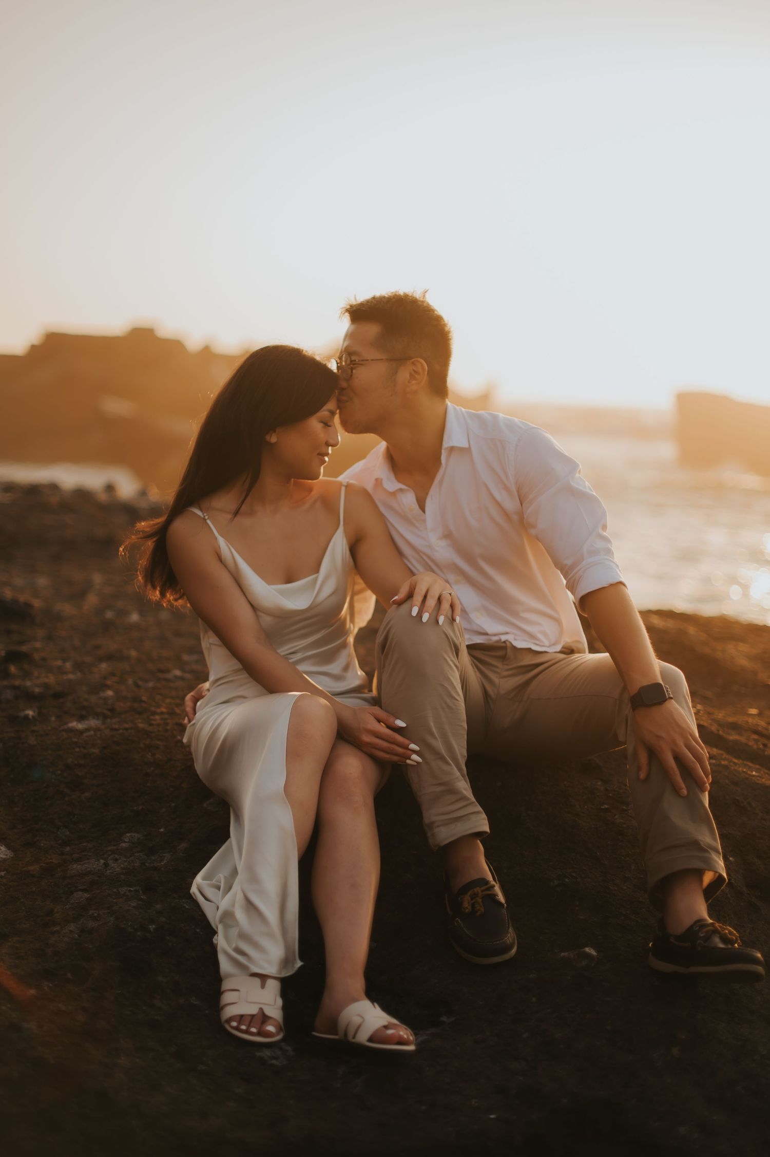 Couple sharing a romantic moment at sunset on rocky beach, woman in white dress, man in white shirt.