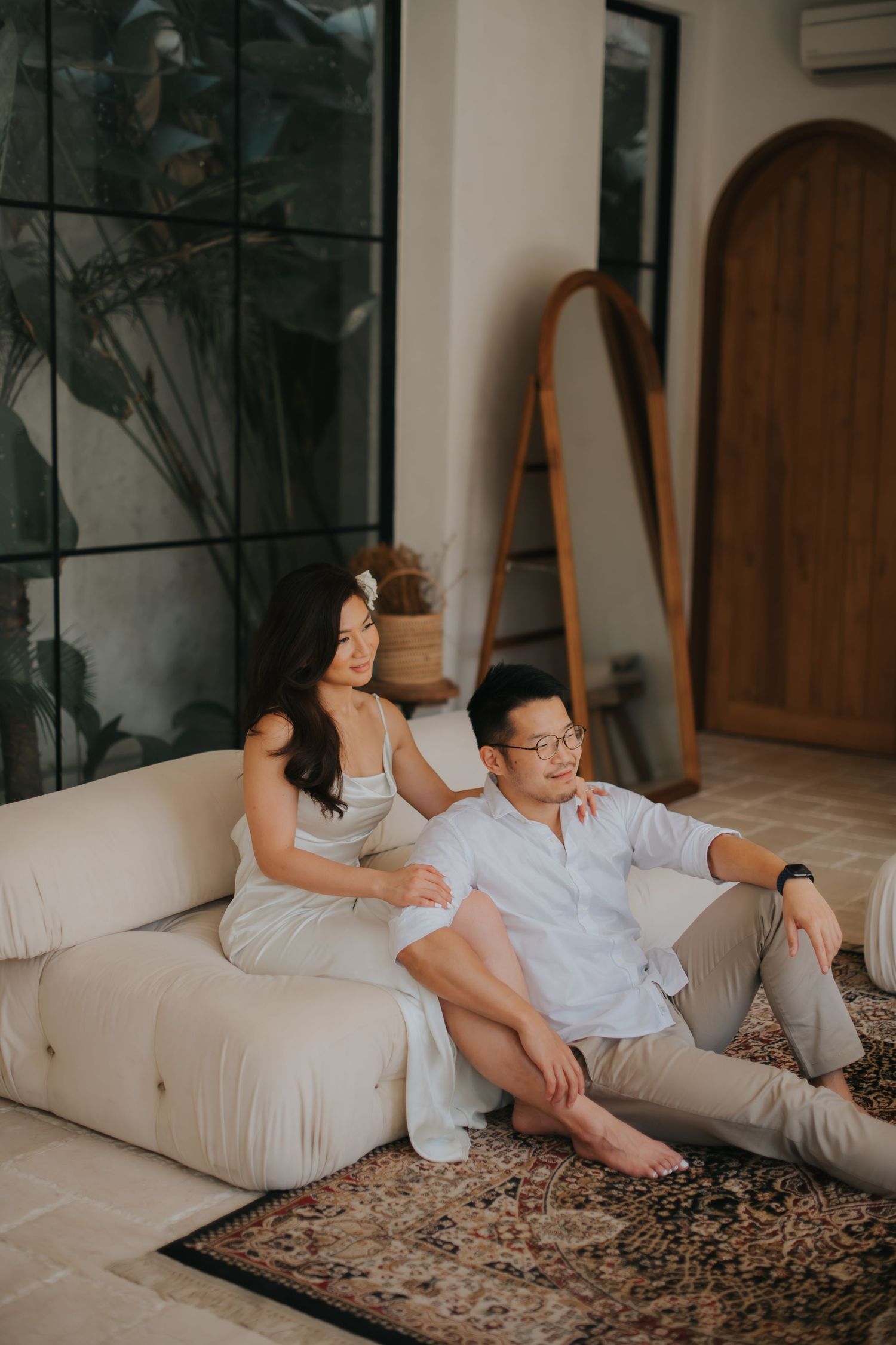 Couple relaxing on a cream sofa in a modern Bali-style living room with arched door and tropical plants.