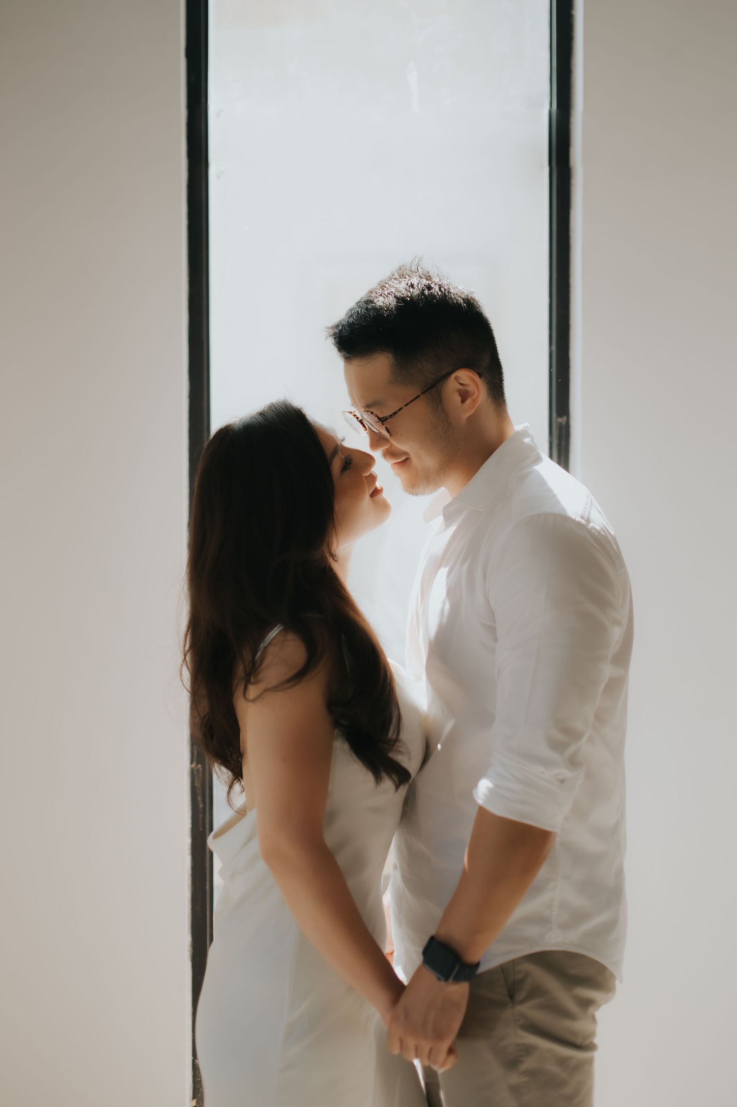 A couple in white outfits shares an intimate moment, holding hands and leaning close together by a bright window.