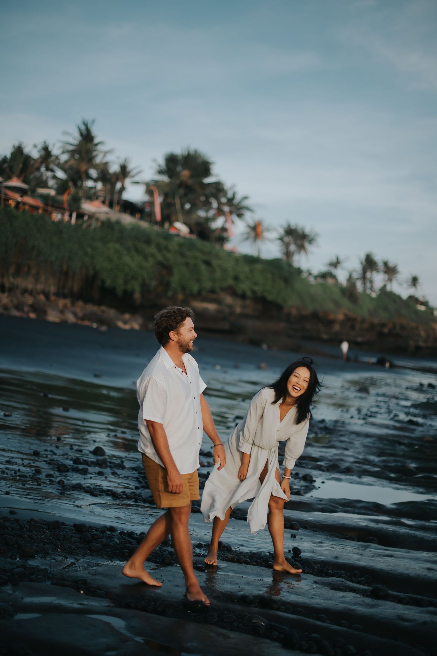 A couple plays together on a black sand beach at sunset with palm trees and rocky coastline in the background.