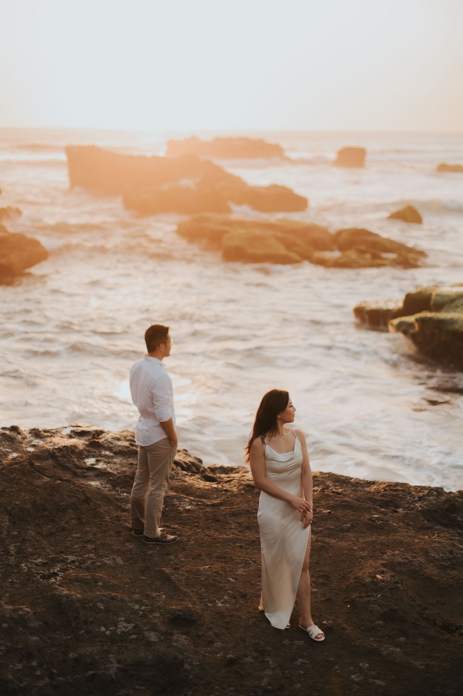 Couple standing on rocky coastal cliffs at golden sunset, woman in white dress, man in white shirt, ocean waves behind.
