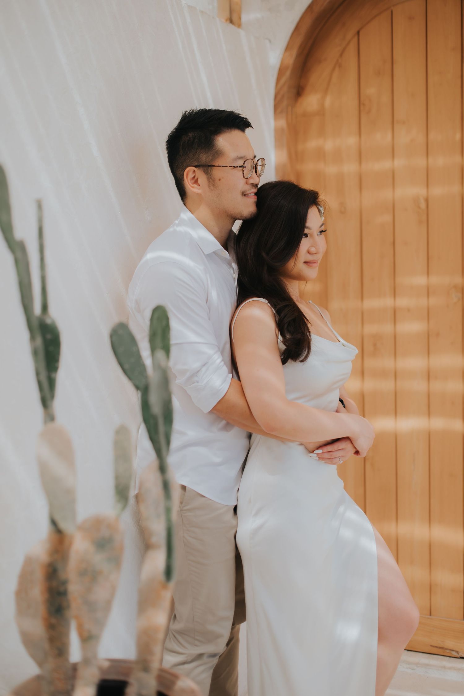 A couple in white outfits embracing near a wooden door and cacti, in a romantic pre-wedding photo shoot.