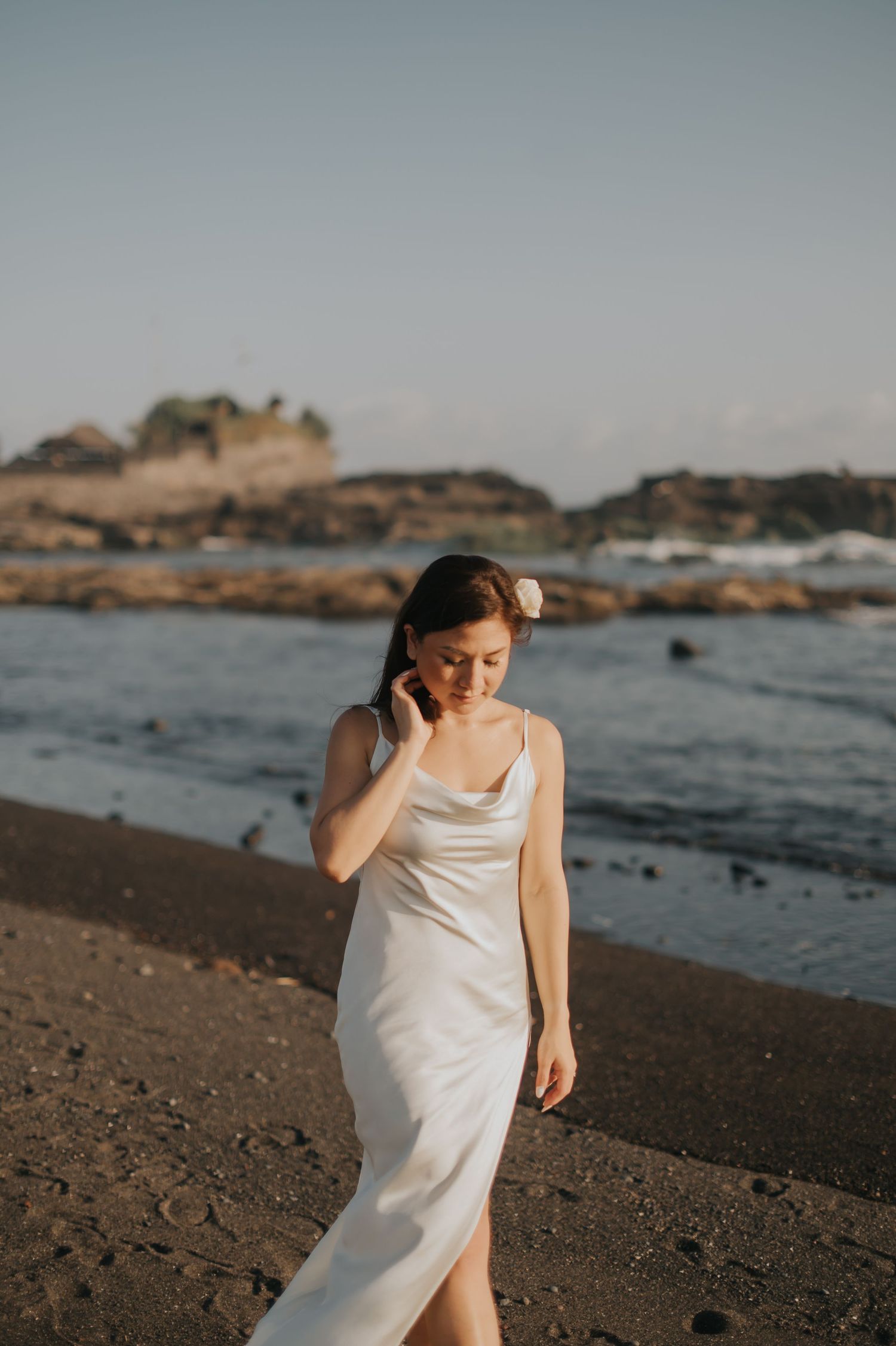 Woman in white satin slip dress walking on a rocky beach at golden hour with ocean waves in the background.