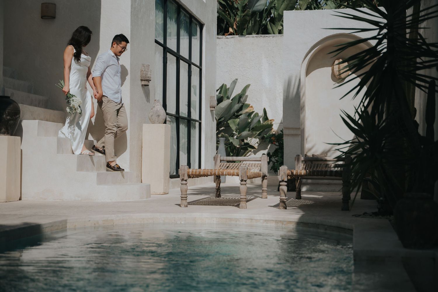 Couple walking down steps beside a serene outdoor pool surrounded by tropical plants and elegant architecture.