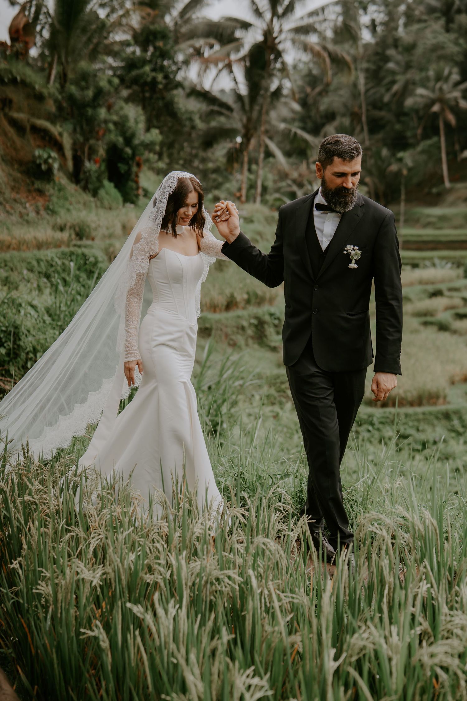 A couple in wedding attire walks through a lush tropical garden with palm trees and tall grass in Bali.