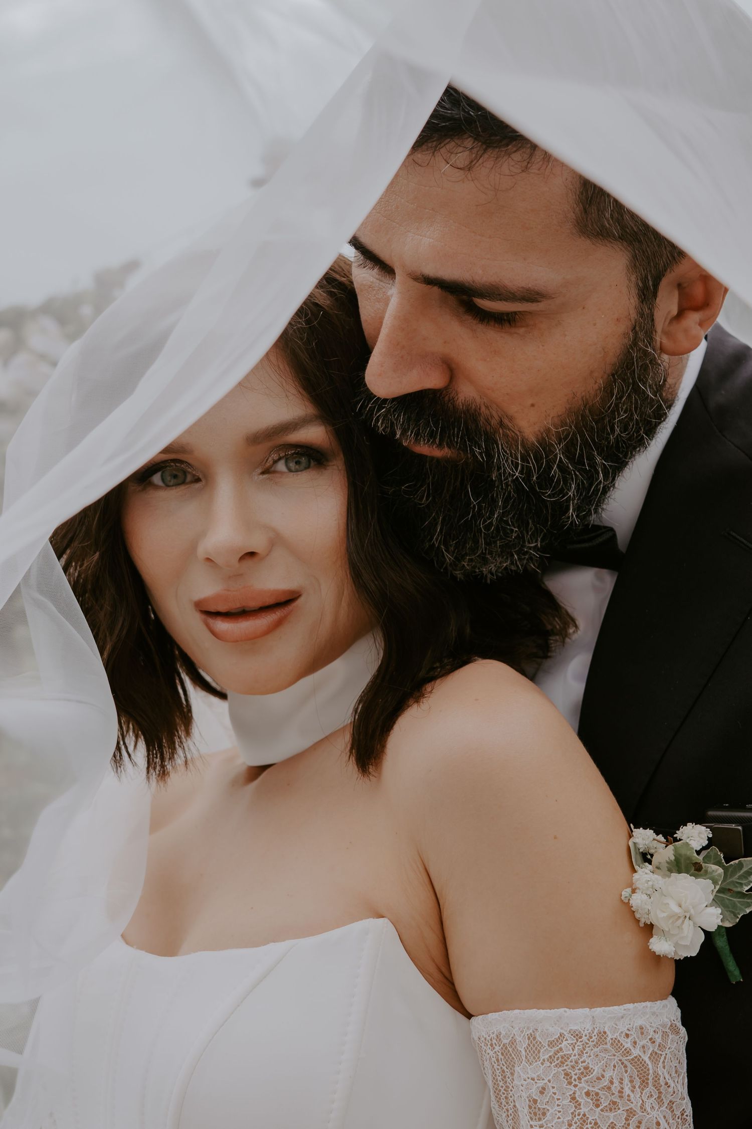 Romantic wedding portrait showing couple under flowing white veil with delicate lace dress and groom in black suit.