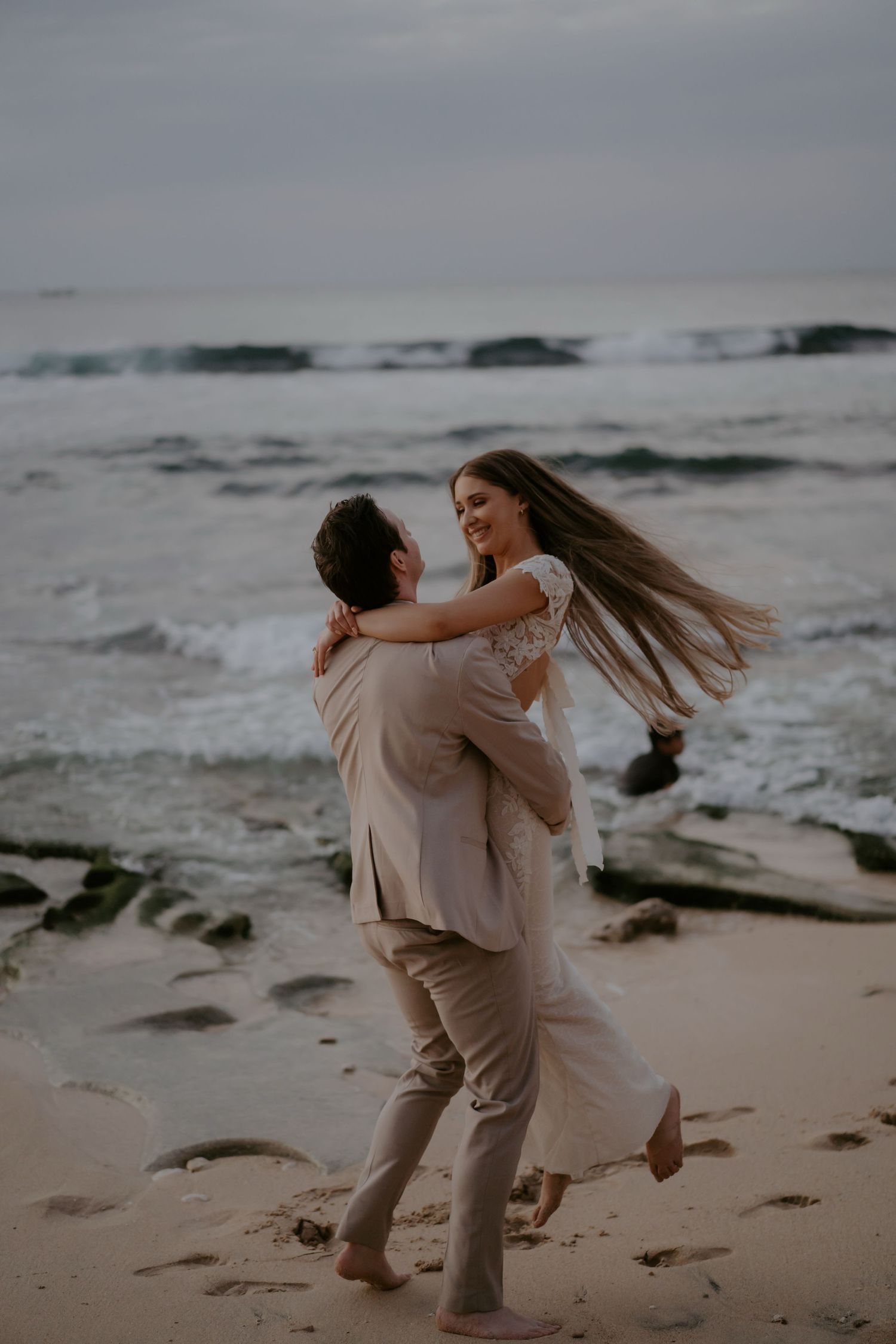A romantic couple embraces and spins on a moody beach at sunset with crashing waves in the background.