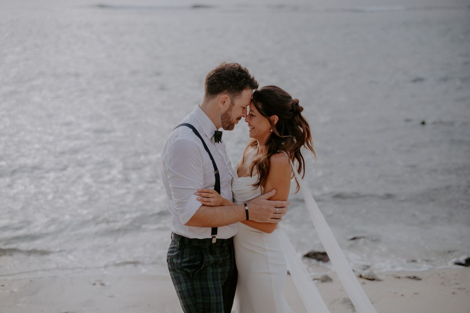 A couple in wedding attire shares a romantic moment on a misty beach with waves crashing behind them.
