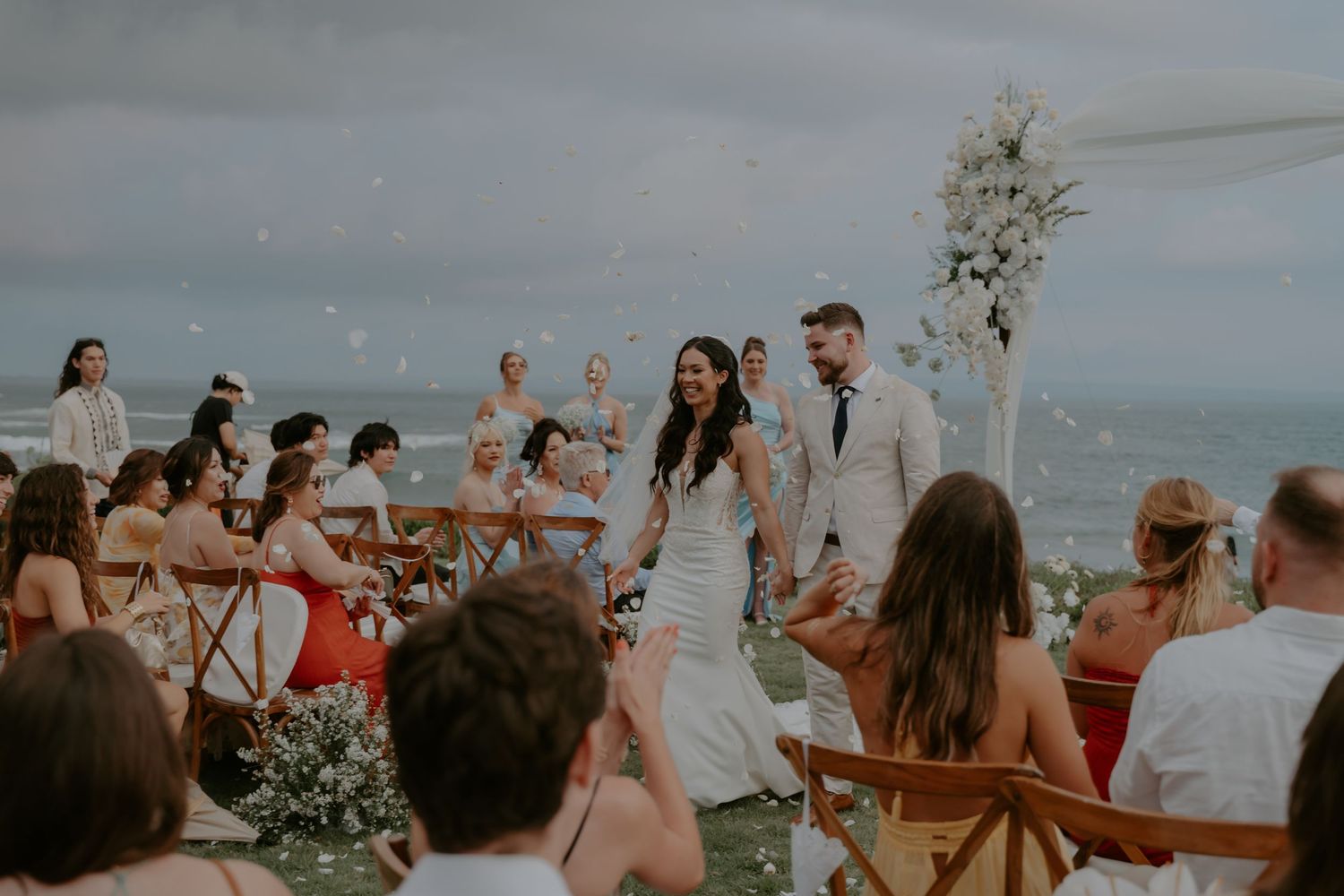 Guests celebrate at a scenic outdoor beach wedding ceremony with wooden chairs and floral decorations at sunset.
