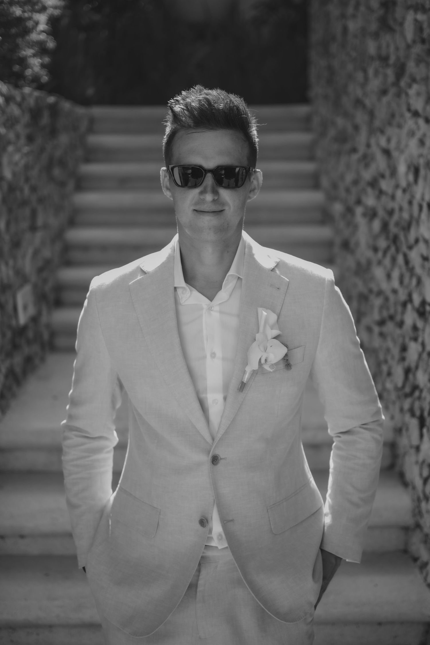 Stylish groom poses in white suit and sunglasses for wedding portraits.