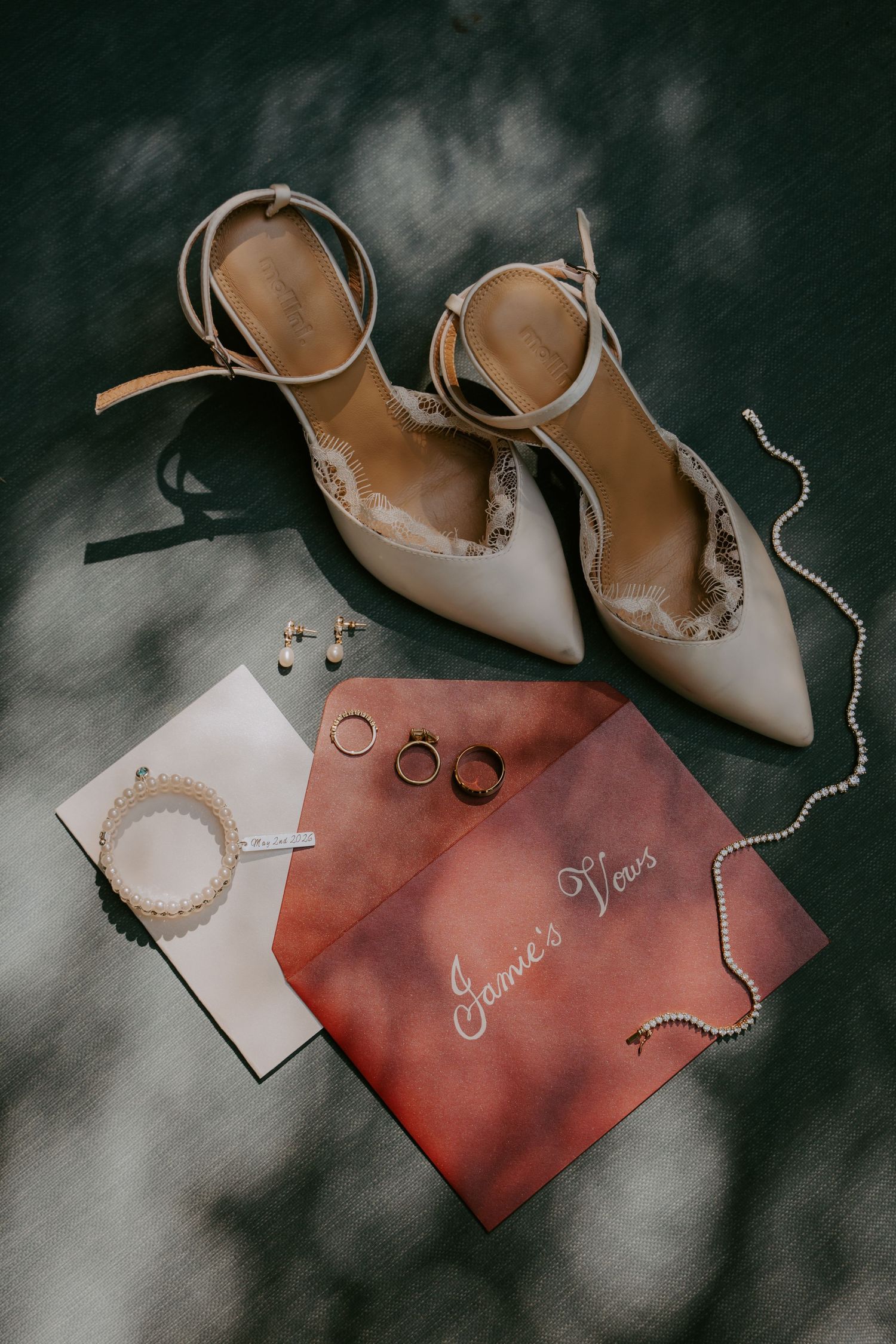 Elegant wedding flat lay featuring nude heels, pearl necklace, gold rings, earrings, and vow booklet on dark surface.