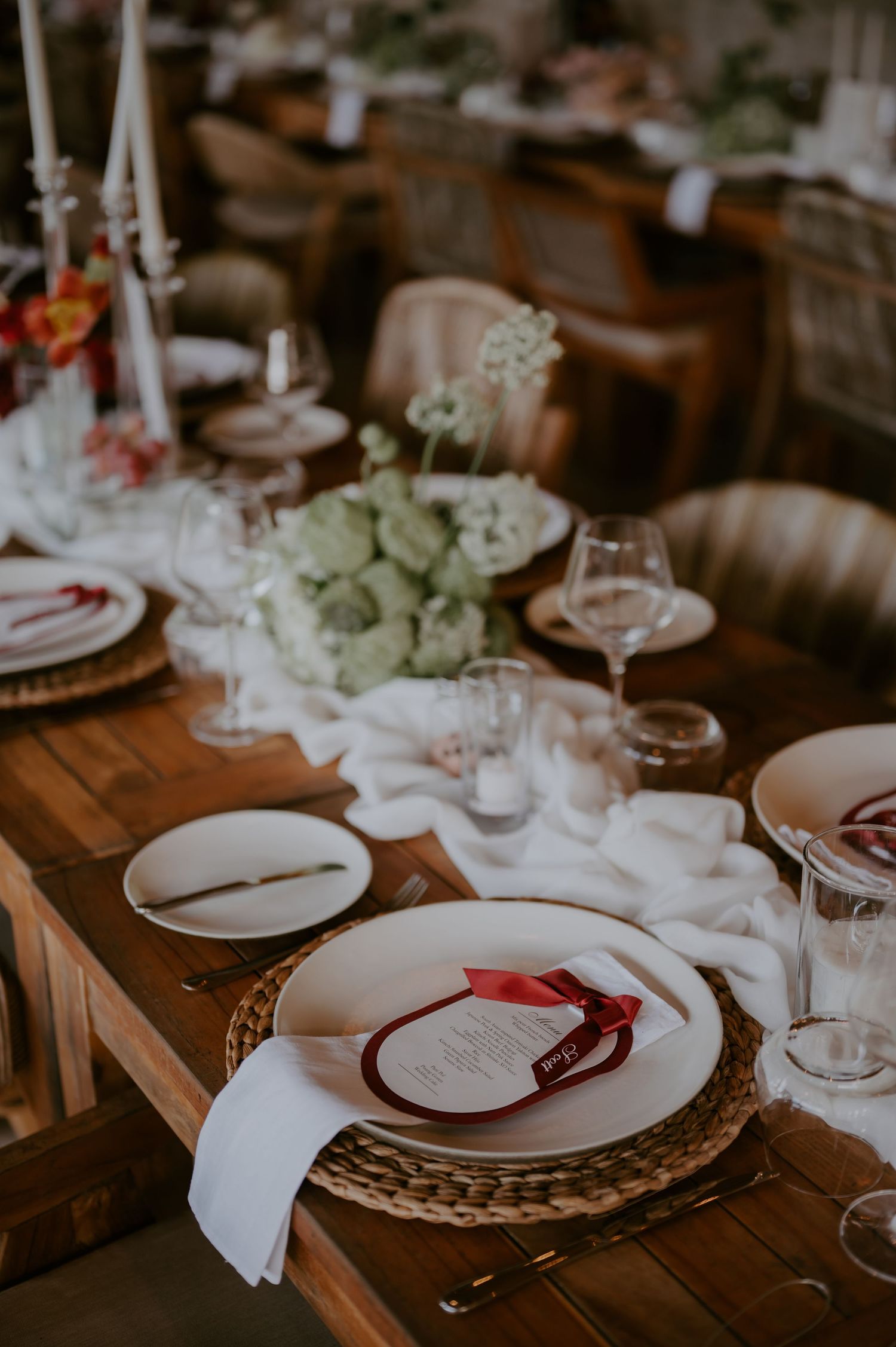 Elegant rustic wedding reception table setting with white linen runner, green floral centerpiece, and red ribbon place cards.