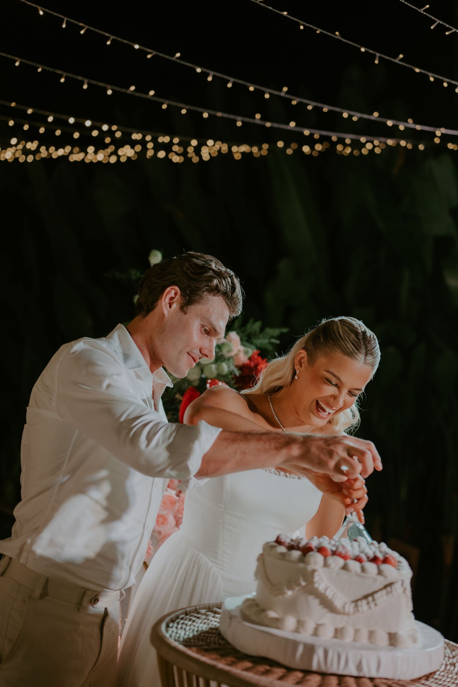 Bride and groom cutting white tiered wedding cake together under string lights at outdoor night reception.