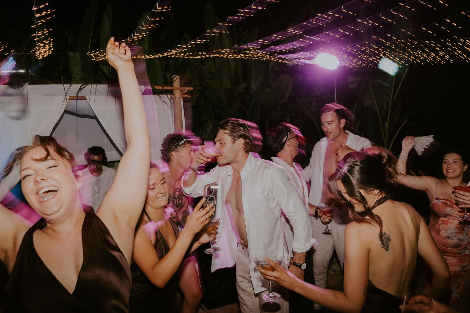 Bride, groom, and wedding guests dancing energetically under string lights at lively outdoor reception.