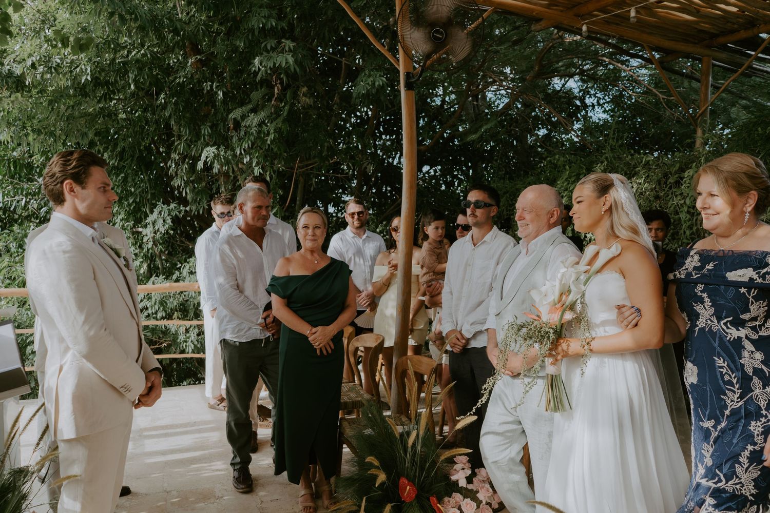 Groom in white suit greets bride and guests during outdoor tropical wedding ceremony.