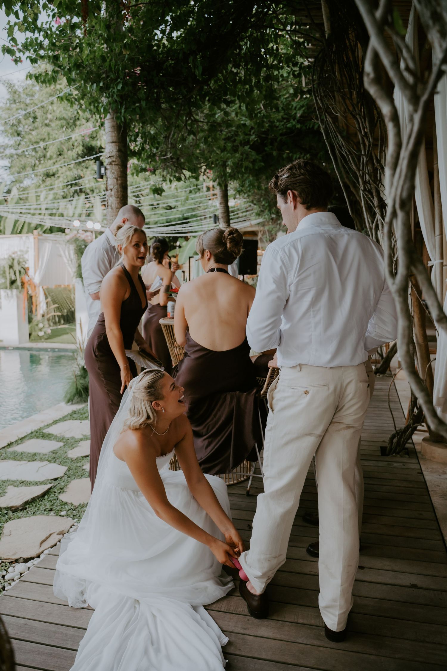 Bride in white gown kneeling to help groom with his shoe on a wooden deck at wedding.