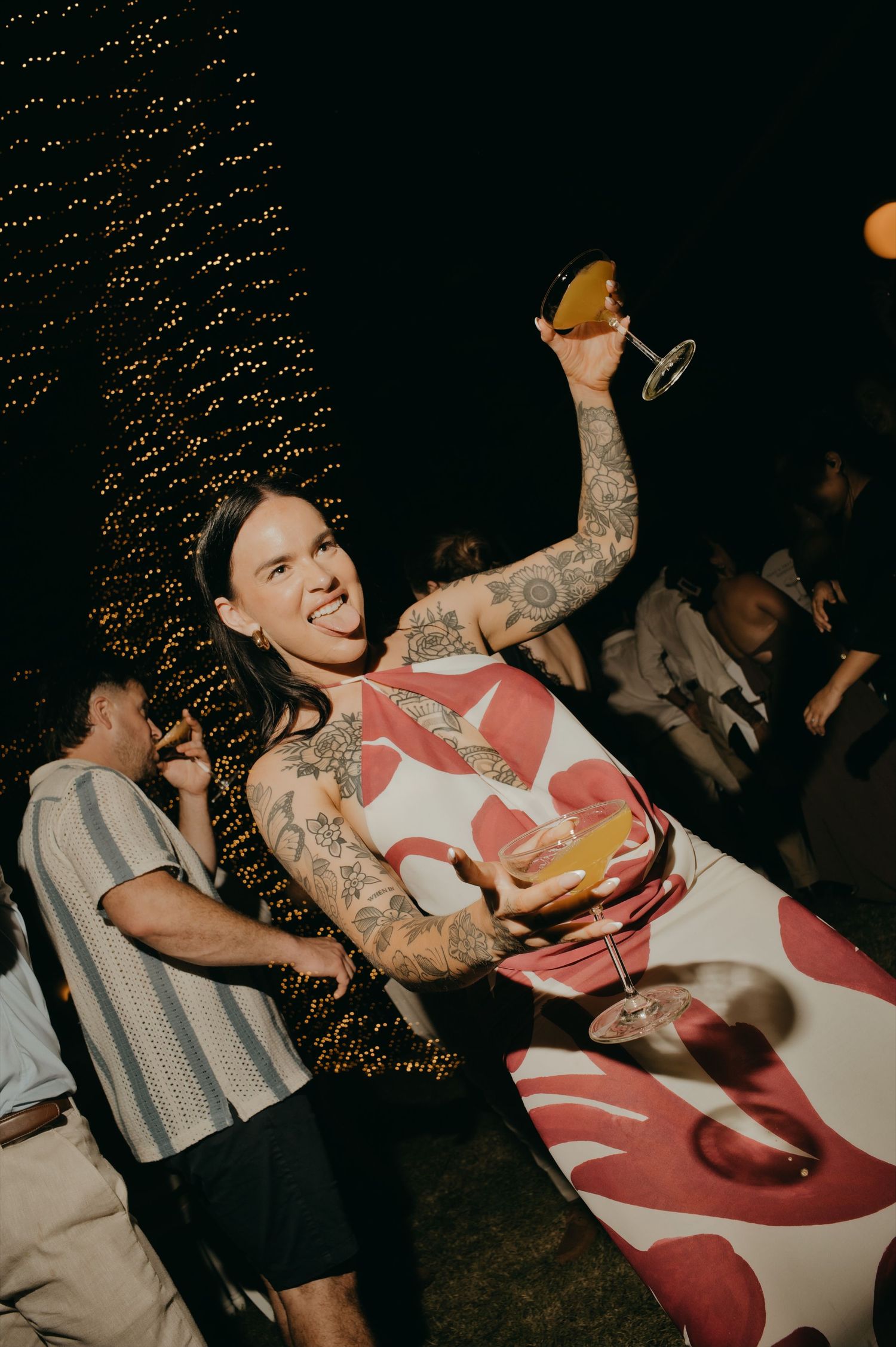 Tattooed wedding guest in floral outfit raises cocktail glass and poses playfully at evening reception.