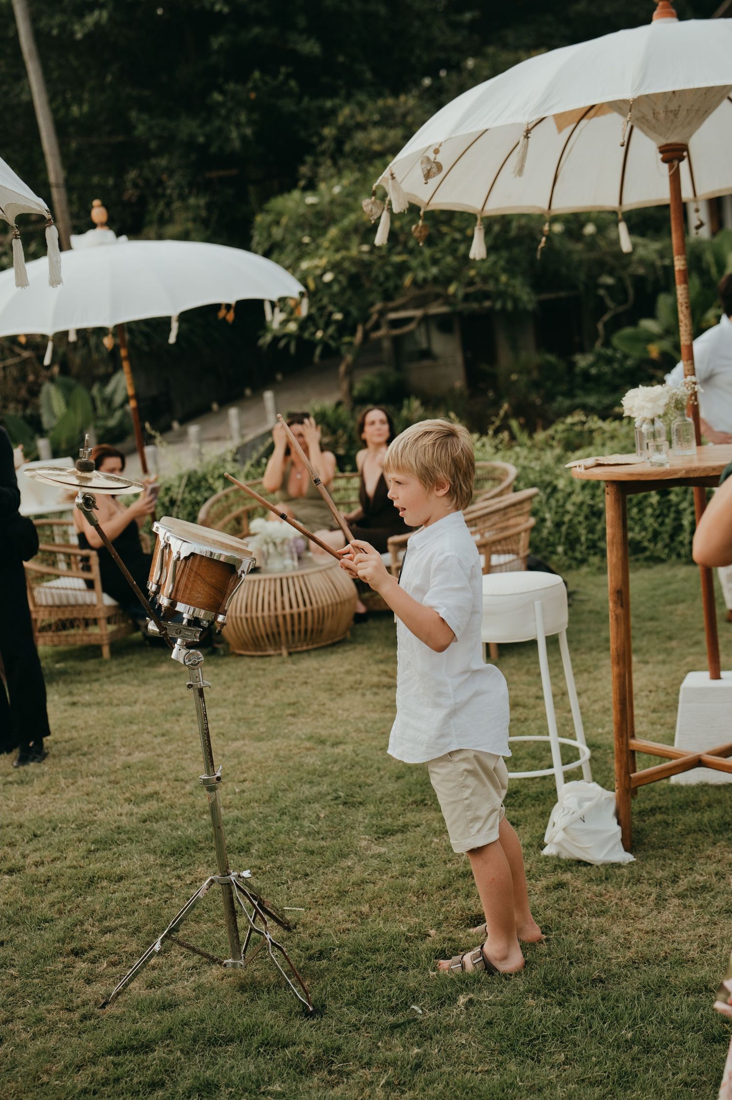 Young boy playing drums at an outdoor wedding reception with white Balinese umbrellas.